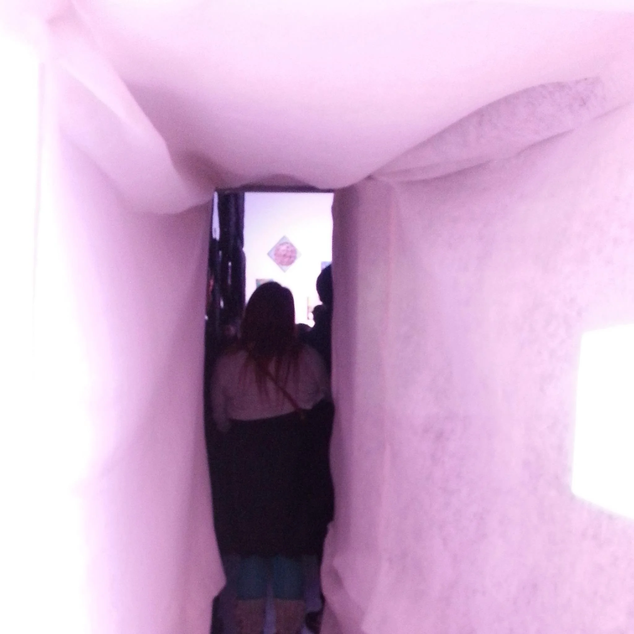 People walking down a narrow, dimly lit indoor hallway with pinkish-purple lighting.