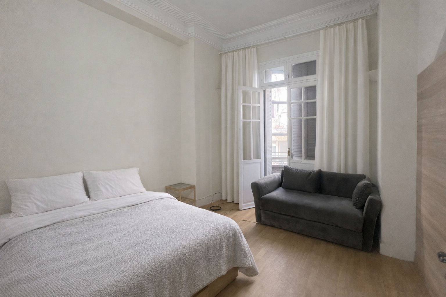 A bedroom with a white bed, a small nightstand, a gray sofa, and large windows with white curtains. Art residency casablanca
