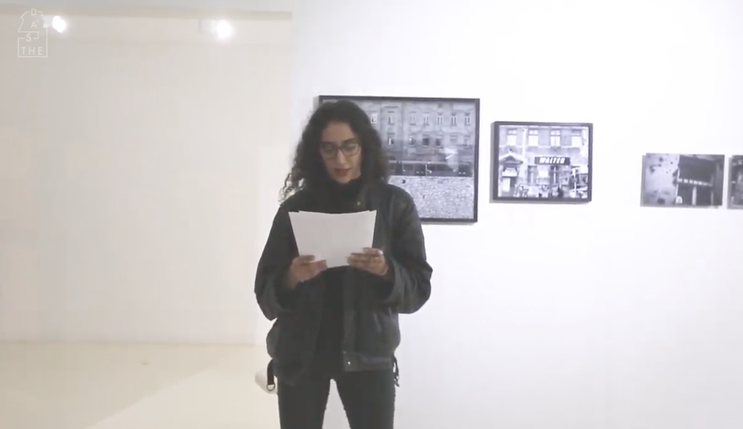 A woman with curly dark hair and glasses standing in an art gallery, reading a piece of paper, with black-and-white photographs hanging on a white wall behind her.