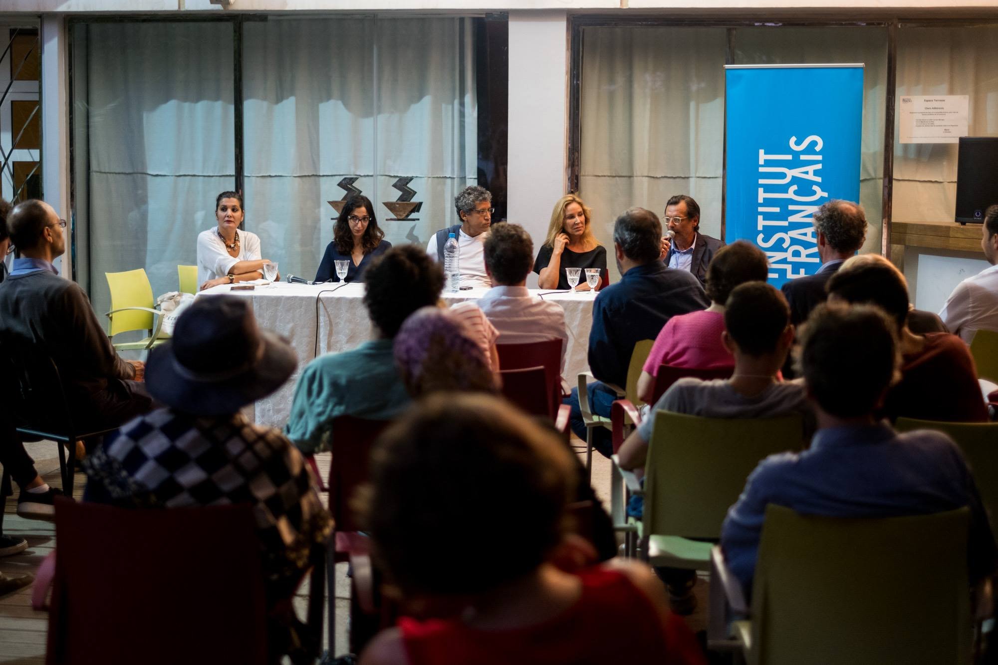 Panel discussion at a conference with six speakers sitting at a long table, audience seated facing the panel, and a blue banner with white text behind them.