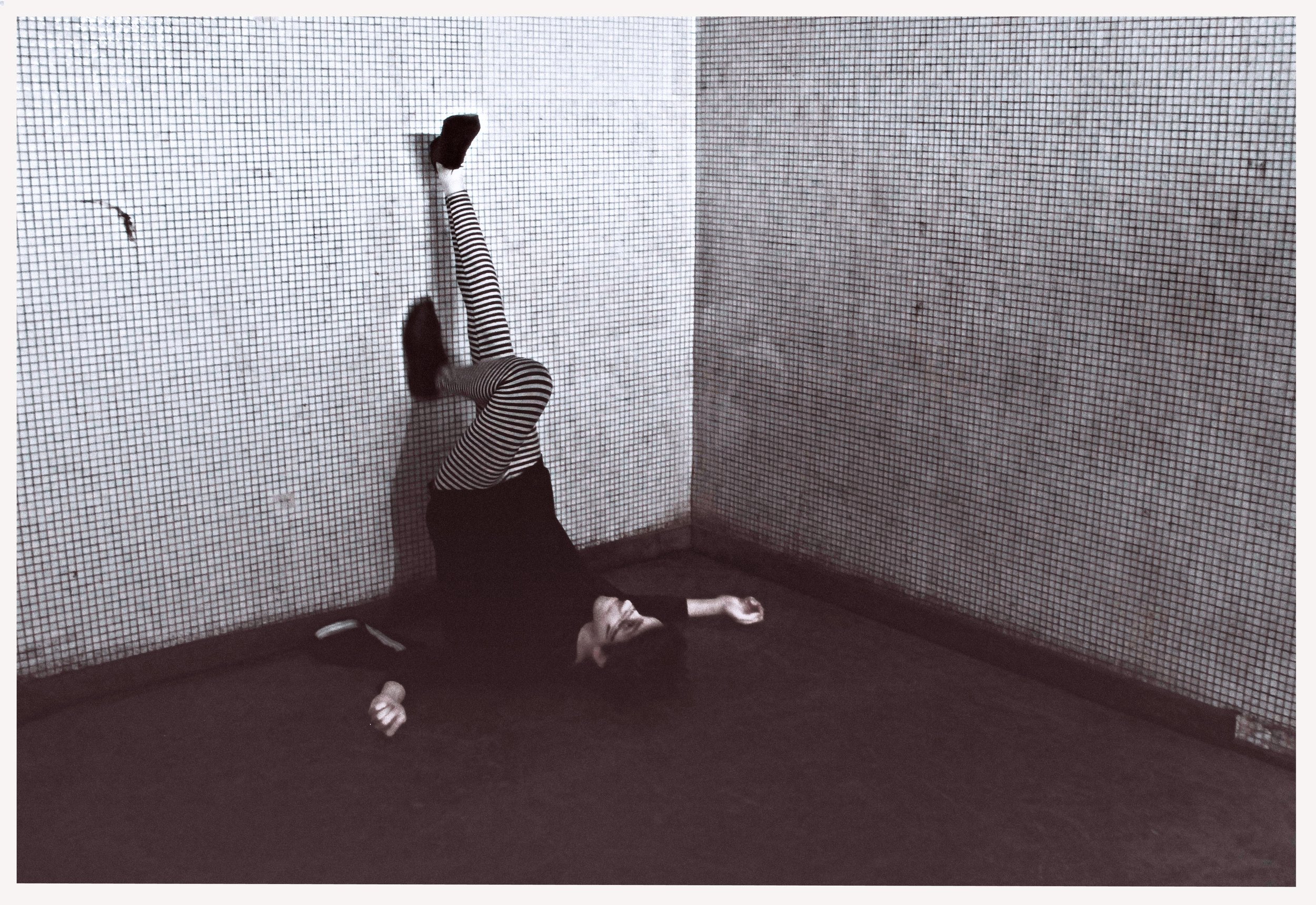 A person lying on their back on a dark floor, with legs extended vertically against a wall with small checkered tiles. The person is wearing striped tights and dark clothing, with a water bottle and phone on the floor nearby.