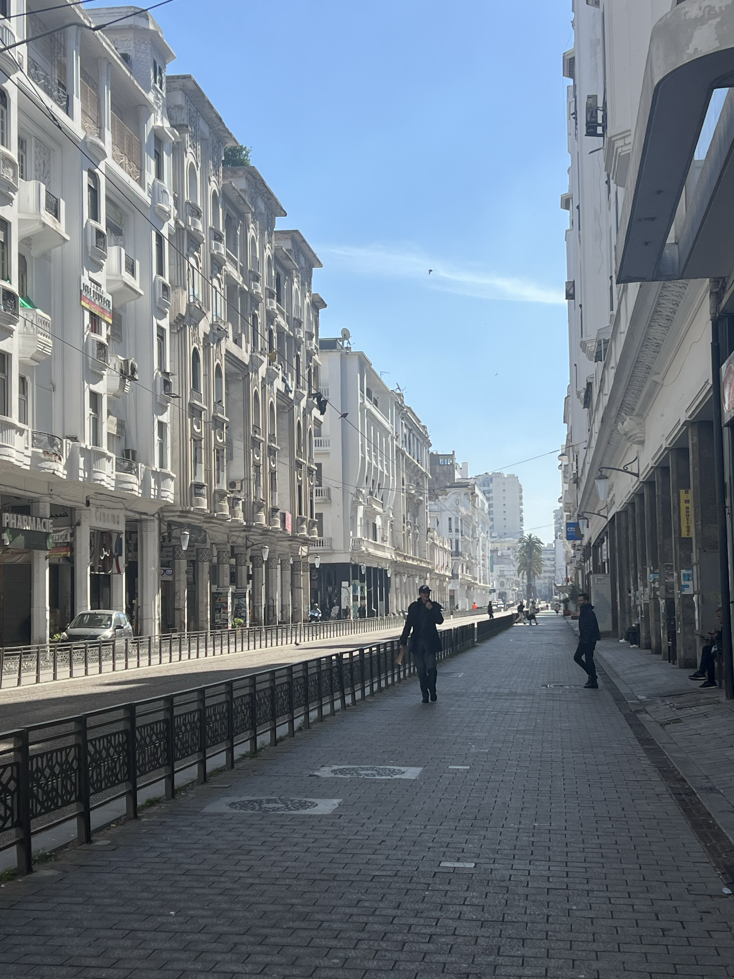 People walking on a city street with white buildings, some with balconies, and a clear blue sky.