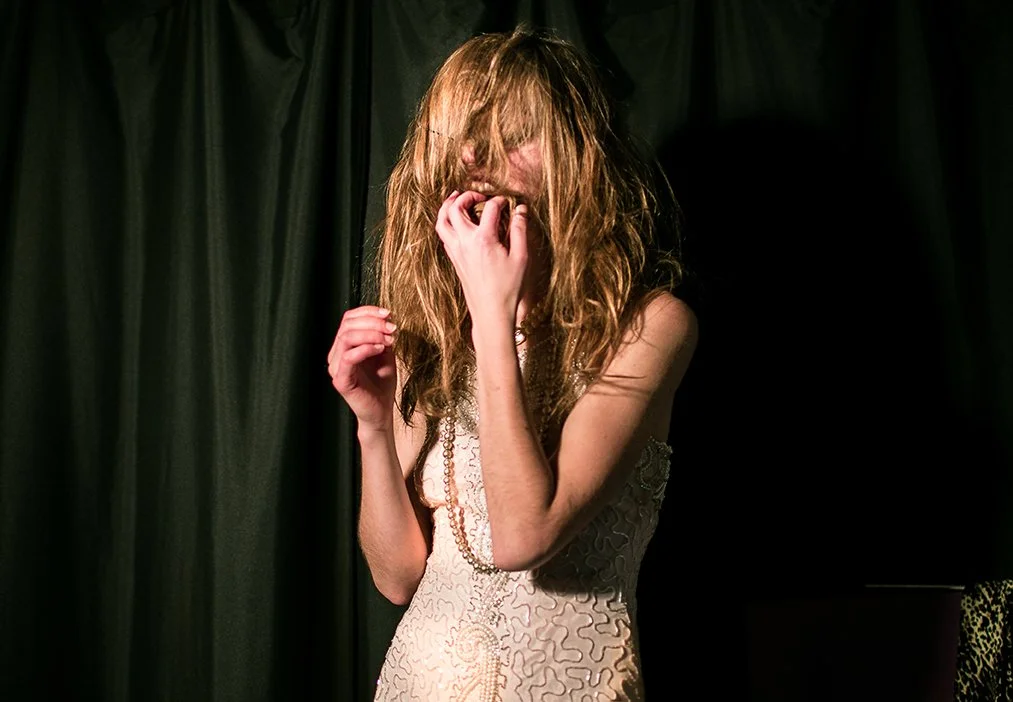 A woman with long, curly red hair covering part of her face, wearing a sleeveless, patterned dress, standing against a dark background with black curtains.