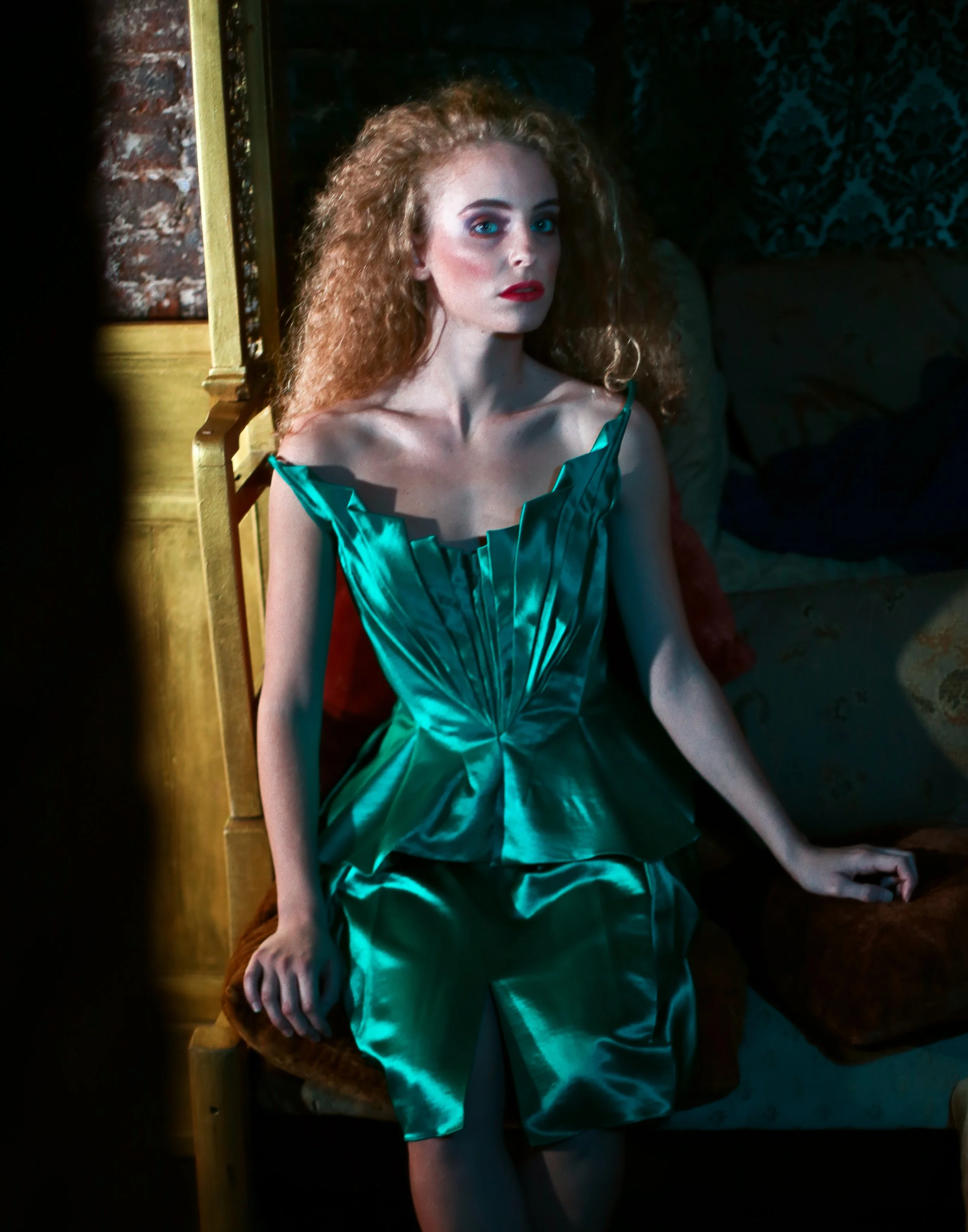 A woman with curly red hair sitting on a wooden bench in a dimly lit room, wearing a shiny aqua green dress.
