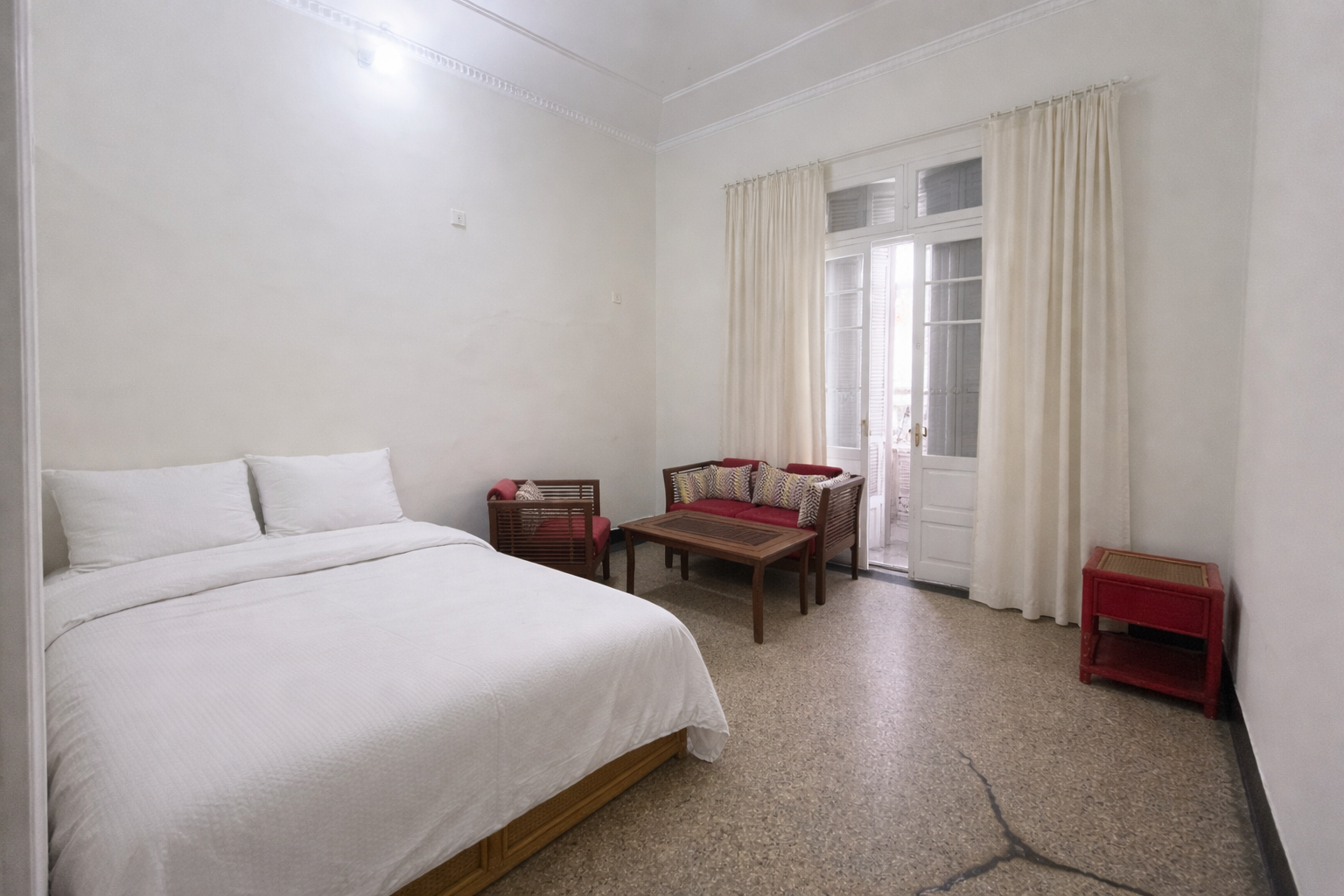 Hotel room with a large bed, white linens, and pillows on the left. In the center, there's a small seating area with a wooden coffee table, a red cushioned sofa, and matching chairs. On the right, cream-colored curtains cover a glass door leading to 