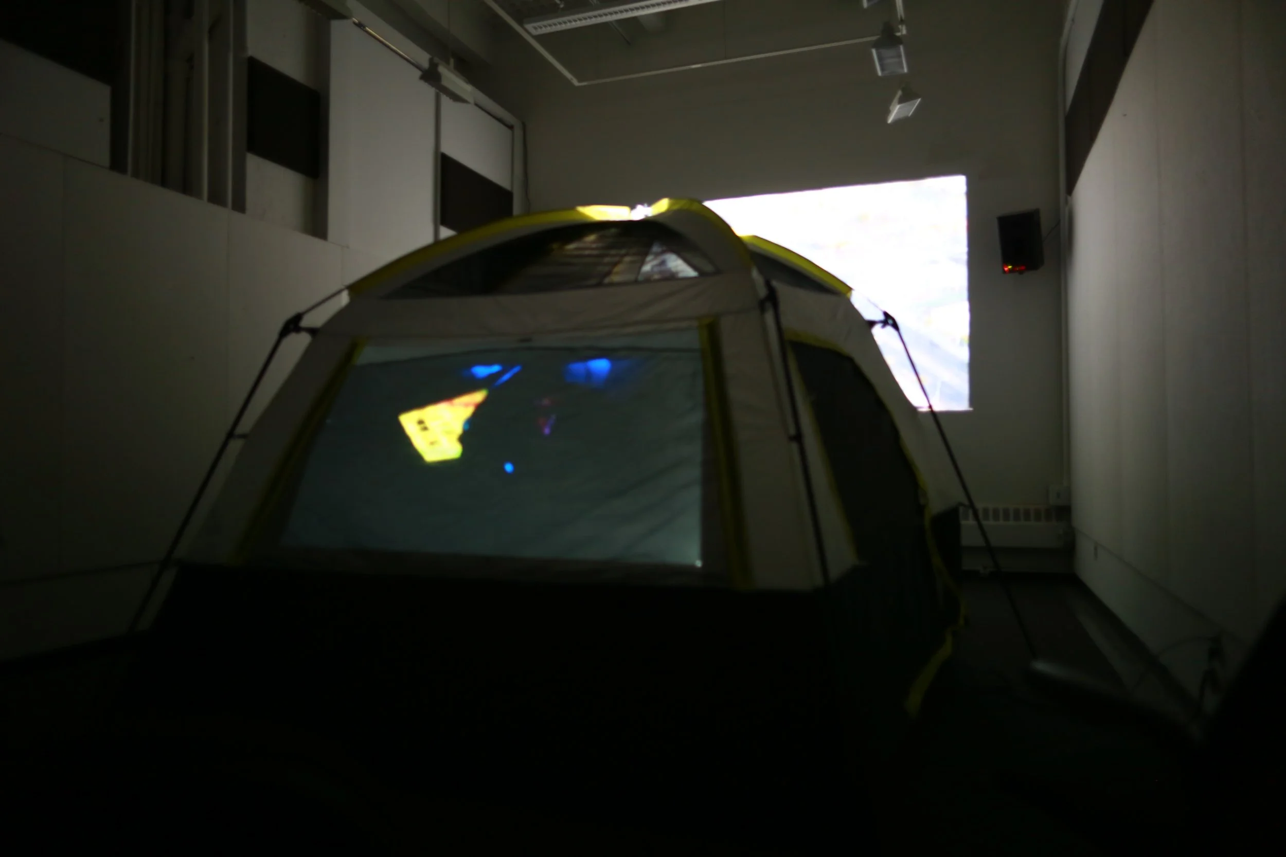 Indoor scene with a yellow and gray camping tent indoors, illuminated by a projection or reflection on its window, against a wall with a bright window above.