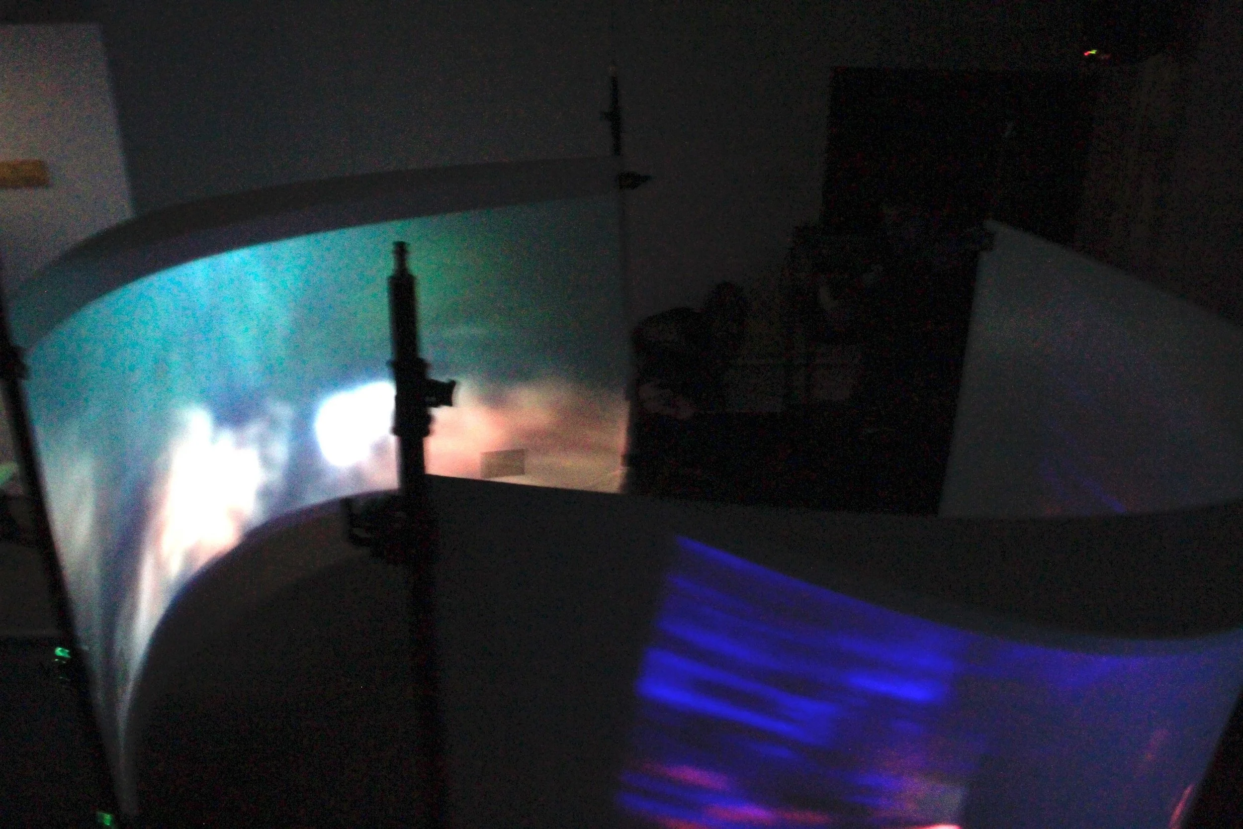 A night scene with a projection of an explosion or fire on a curved surface, with a silhouette of a gun or rifle in the foreground and dark indoor surroundings.