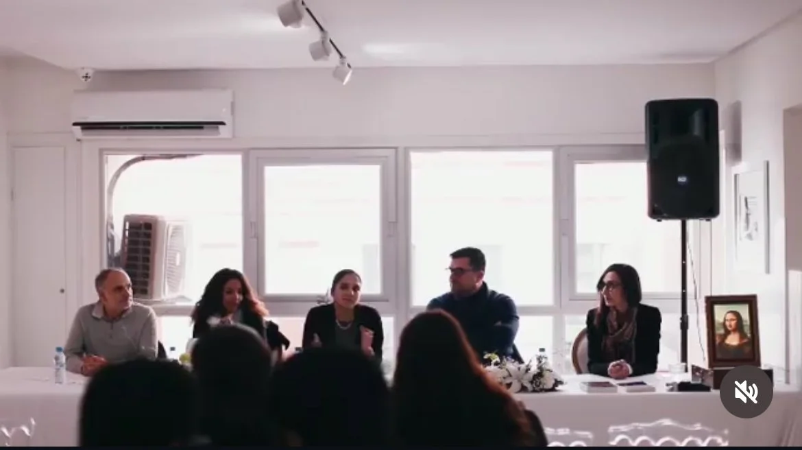 A panel of five people sitting at a long table during a conference or meeting, with a large window behind them, a speaker, and framed photograph on the right side.