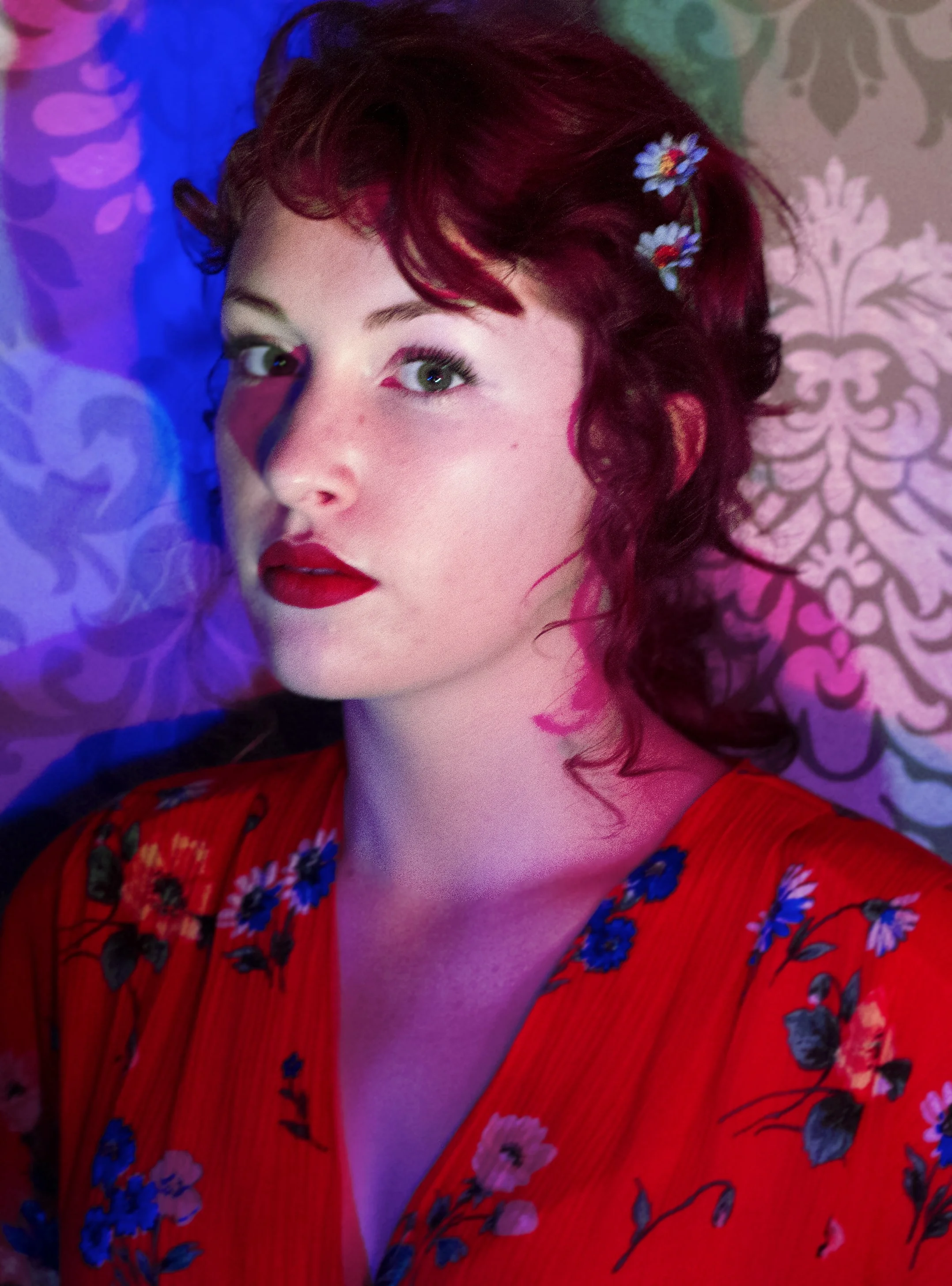A woman with short curly dark red hair and fair skin, wearing a red floral dress, with small flowers in her hair. She has makeup with red lipstick and green eyes, and is illuminated by colorful blue, purple, and pink lighting against a patterned background.