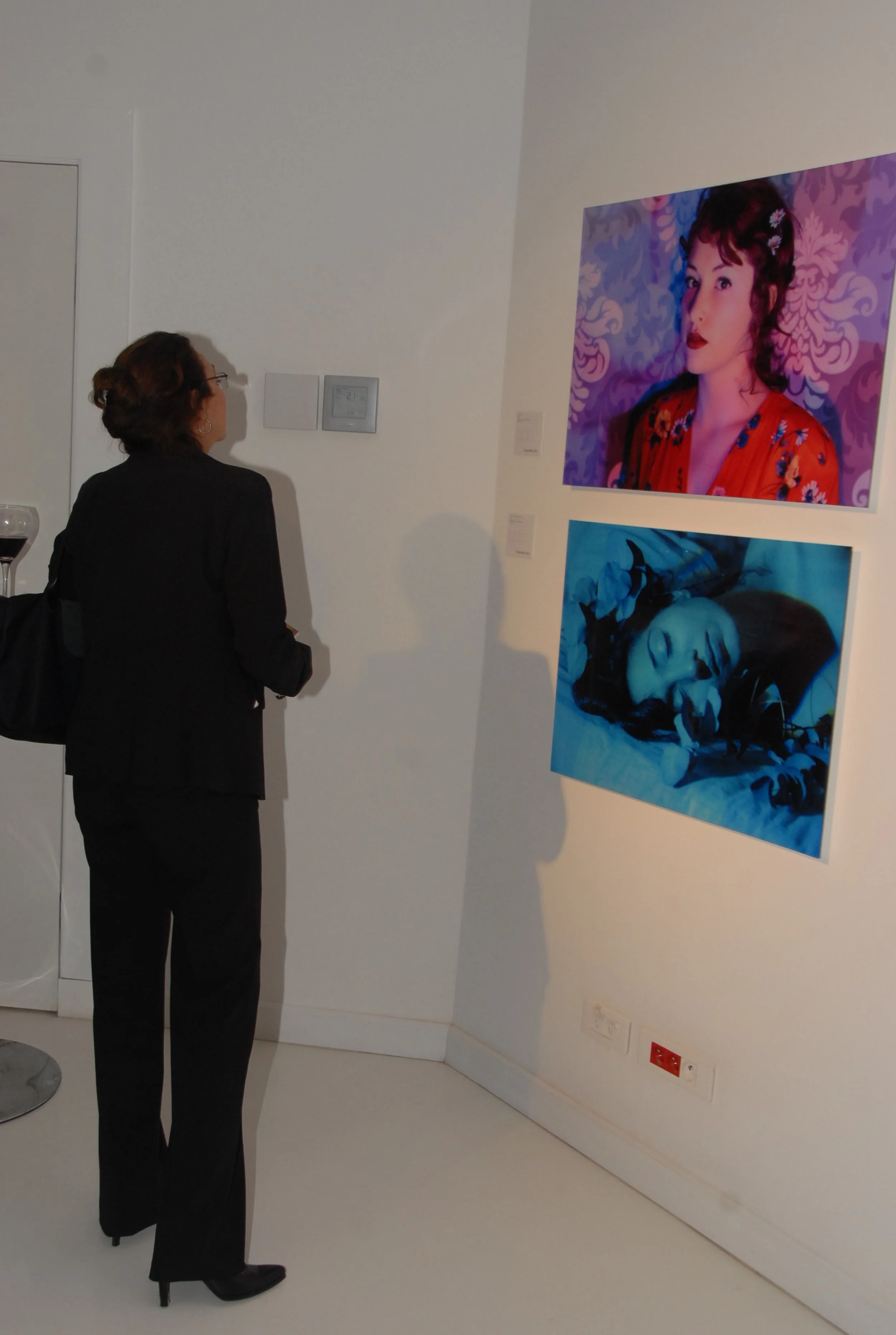 A woman in a black suit with glasses observing two portrait photographs on a white wall of an art gallery.