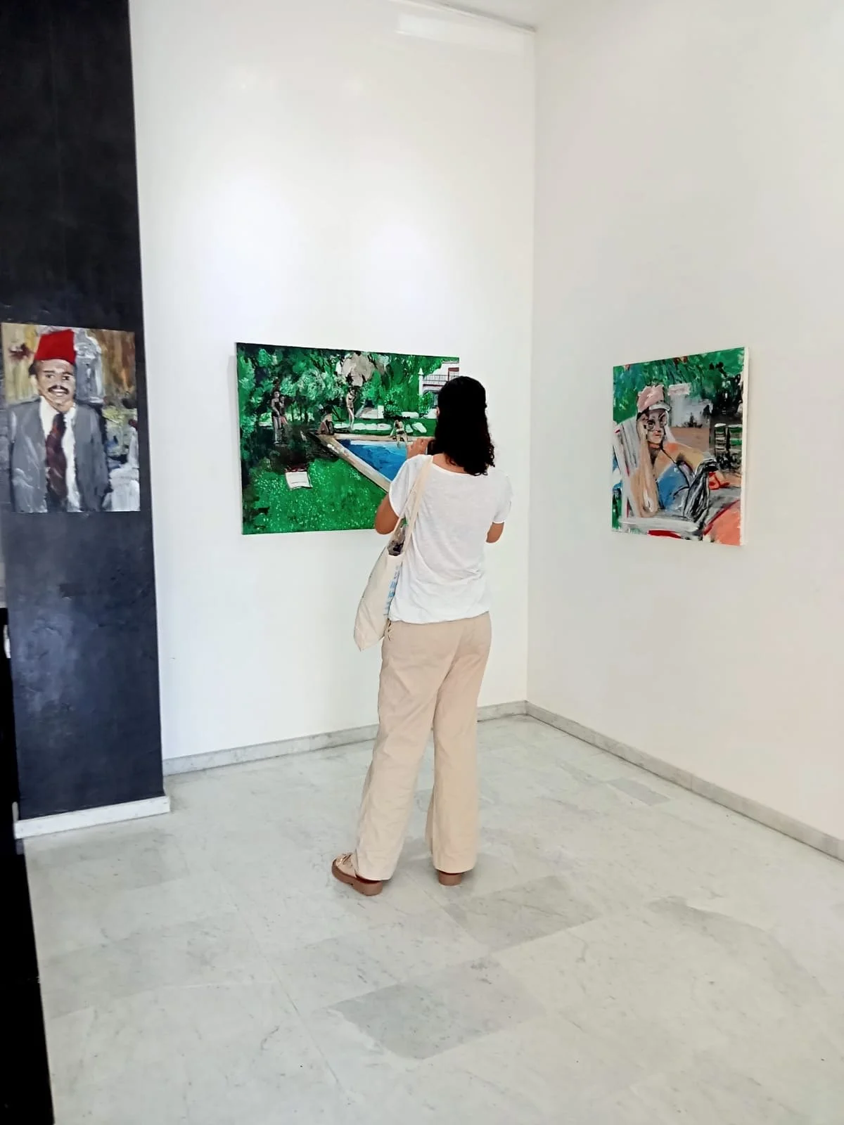 A woman standing in an art gallery, observing two paintings on the white walls, with a third painting on a black panel to the left.