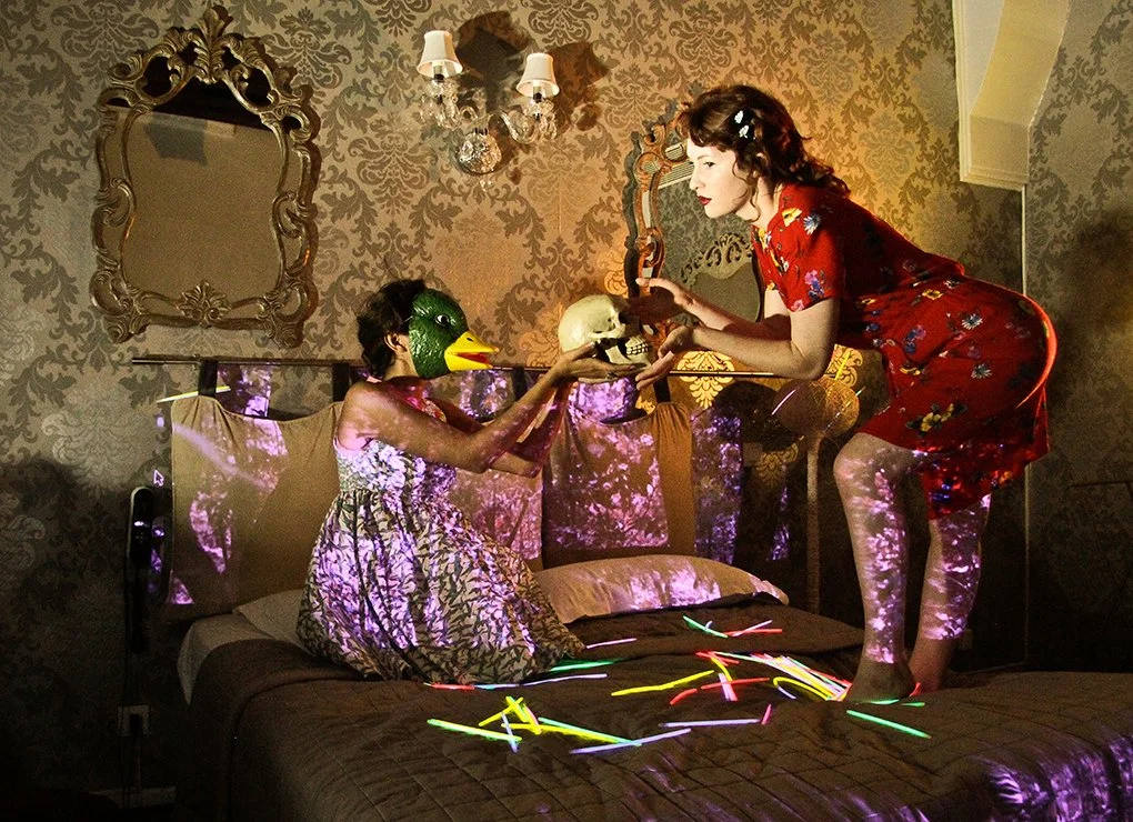 Two women in vintage-style clothing inside a decorated bedroom, one wearing a duck mask and the other holding a skull, with neon-colored lights scattered on the bed.