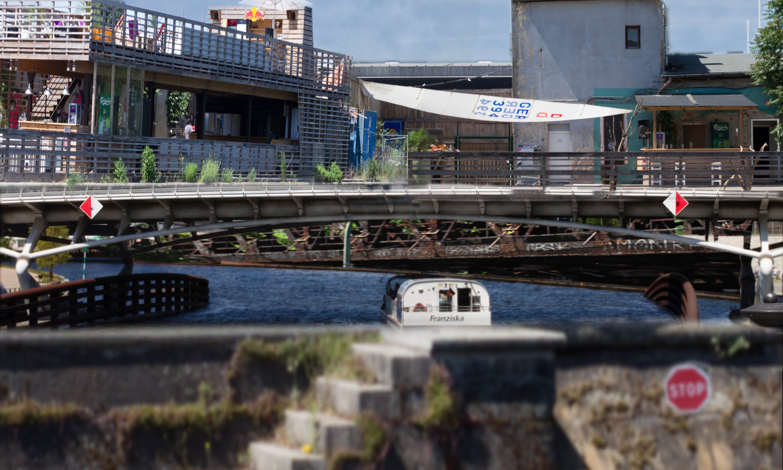 A city scene with a canal, a boat named Franziska on the water, and modern buildings with outdoor terraces and greenery above the canal.