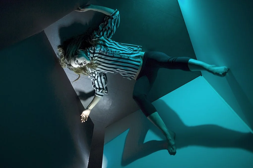 A woman lying on the floor in a modern, geometric room with blue lighting, wearing a striped shirt and black pants.
