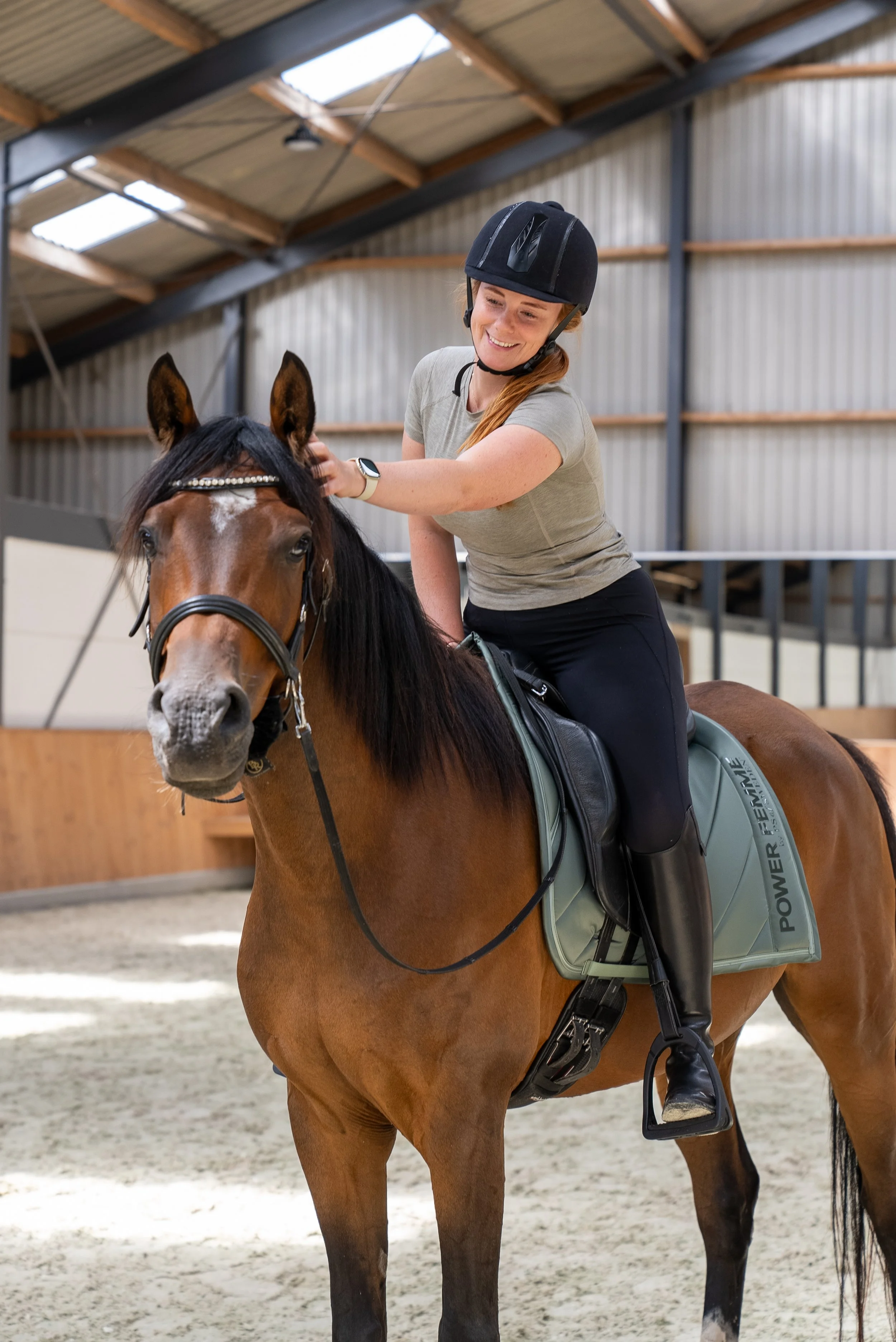 A woman riding a horse in an indoor riding arena, smiling and petting the horse's head.