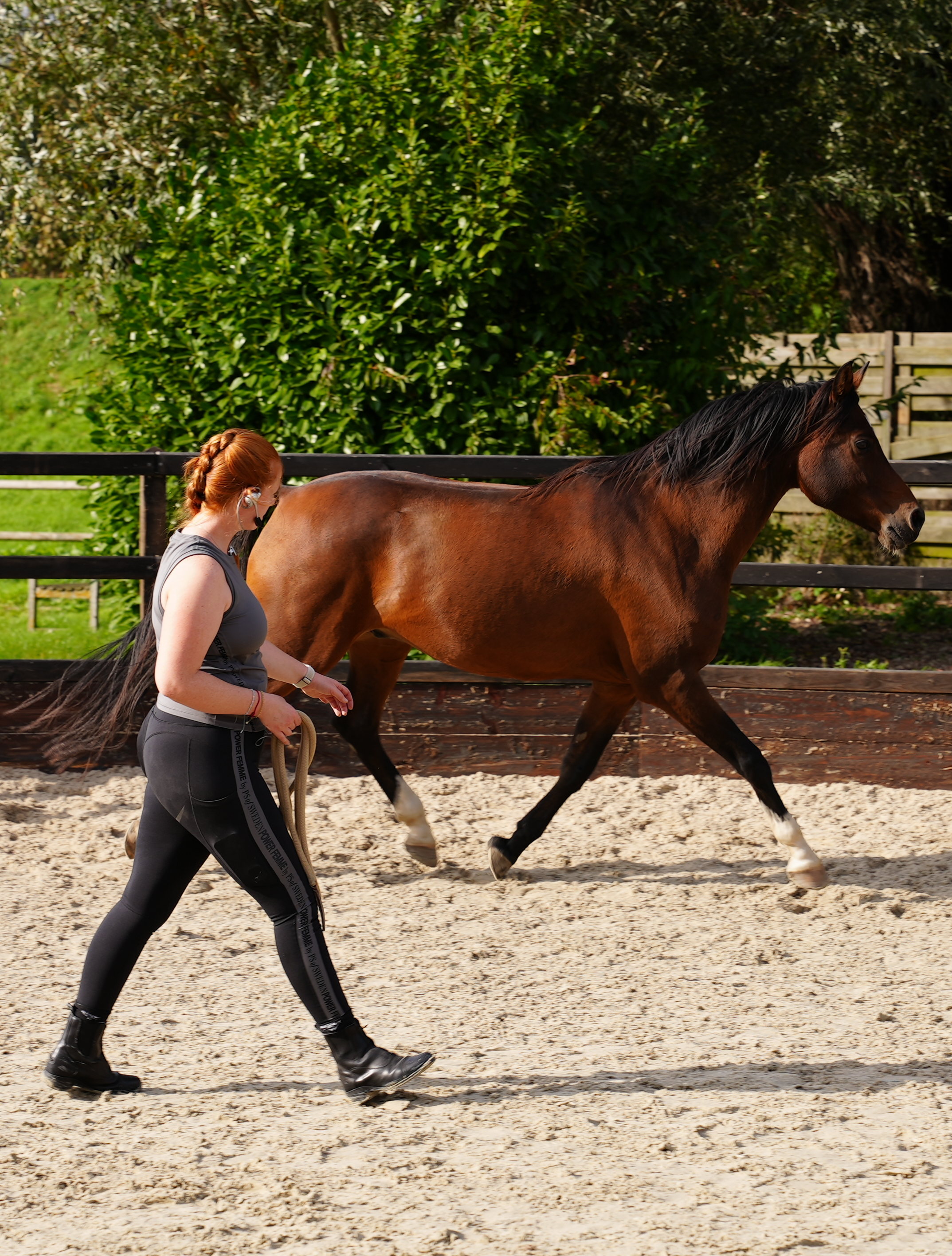 A woman with red hair, wearing a sleeveless top, black leggings, and black boots, is leading a brown horse in an outdoor riding arena with a sand surface, surrounded by a wooden and black fence, with green bushes and trees in the background.