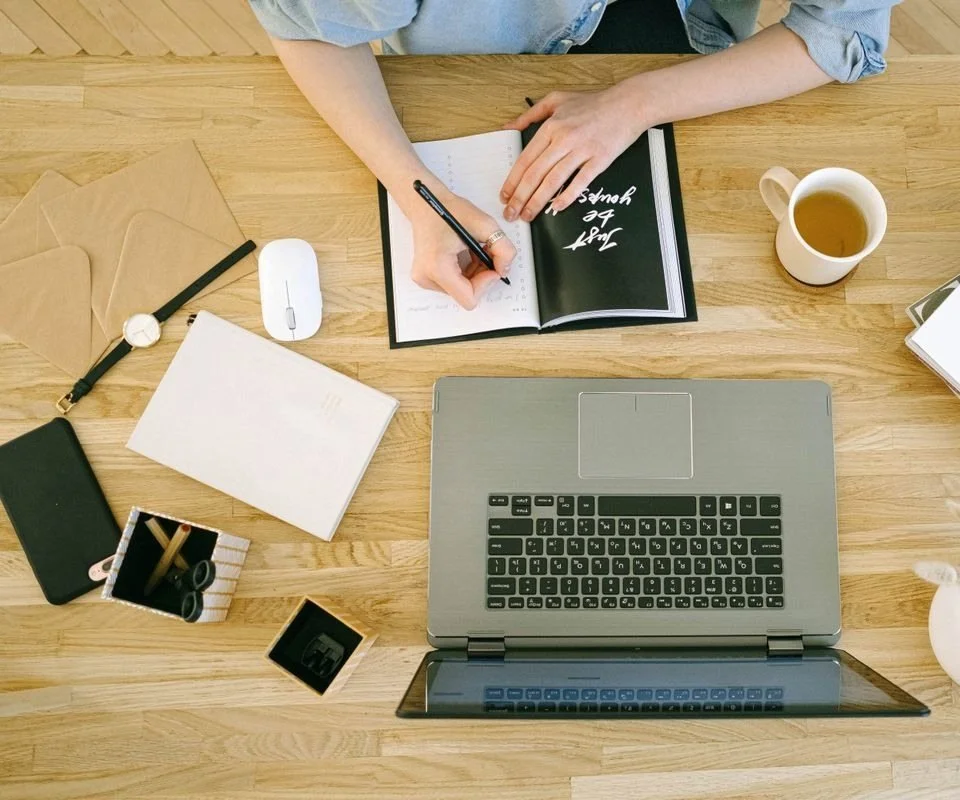 An overhead view of a hand writing in a notebook beside an open laptop and a cup of tea on a wooden desk.