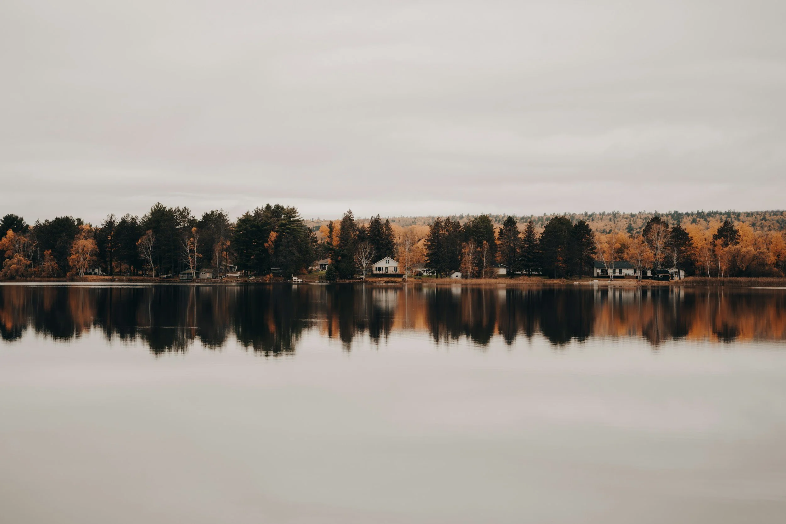 Calm lake reflection of trees and houses along the shoreline with overcast sky.