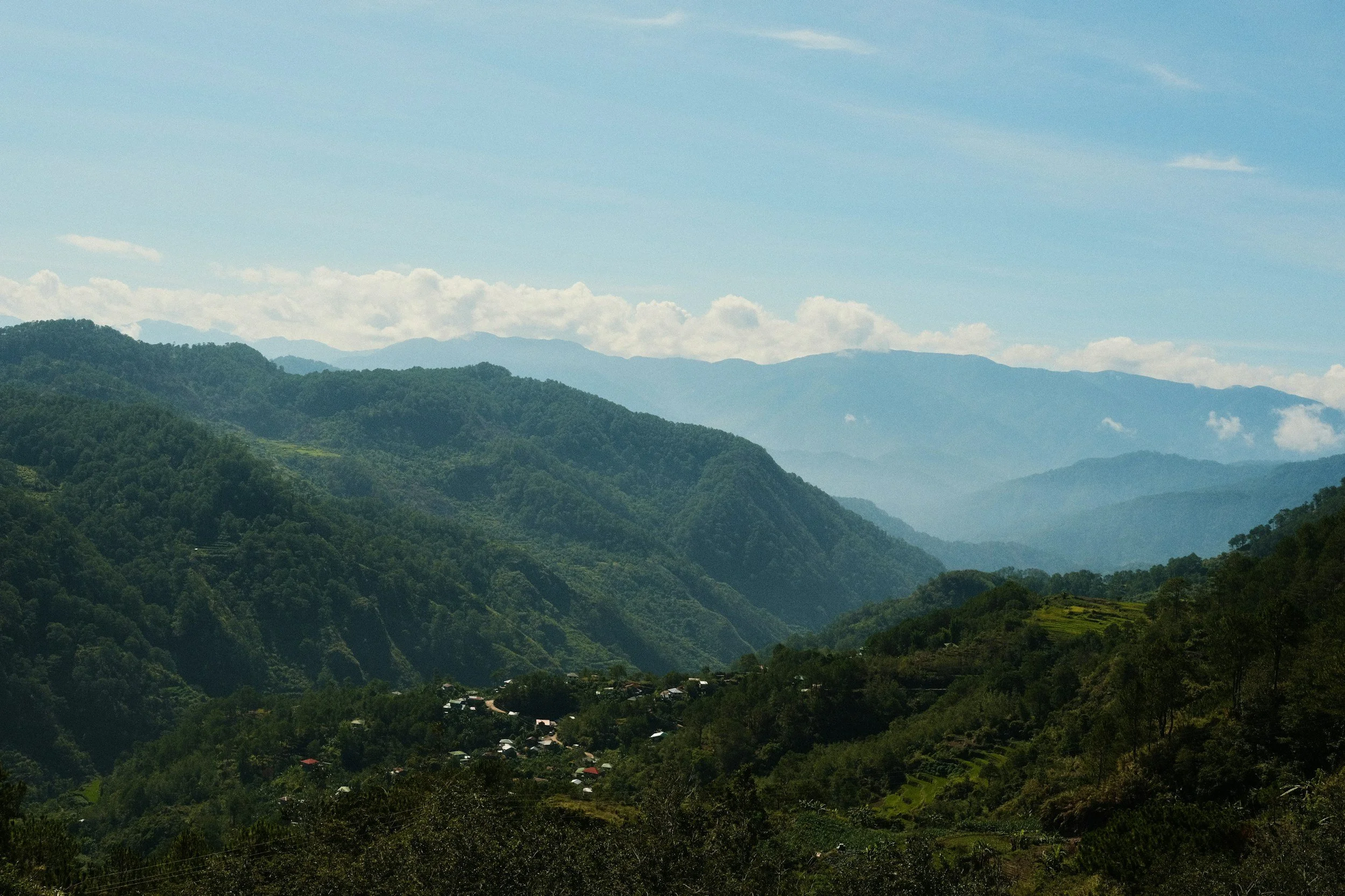 Scenic view of lush green mountains with small village houses scattered on the hillside beneath a mostly clear sky with some clouds.