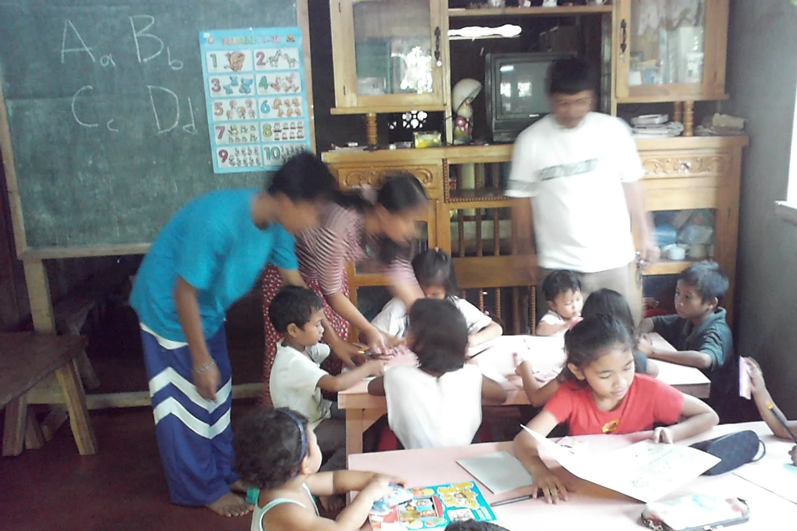 A classroom with children sitting at tables learning, with teachers assisting. There is a chalkboard with alphabet and number posters on the wall.