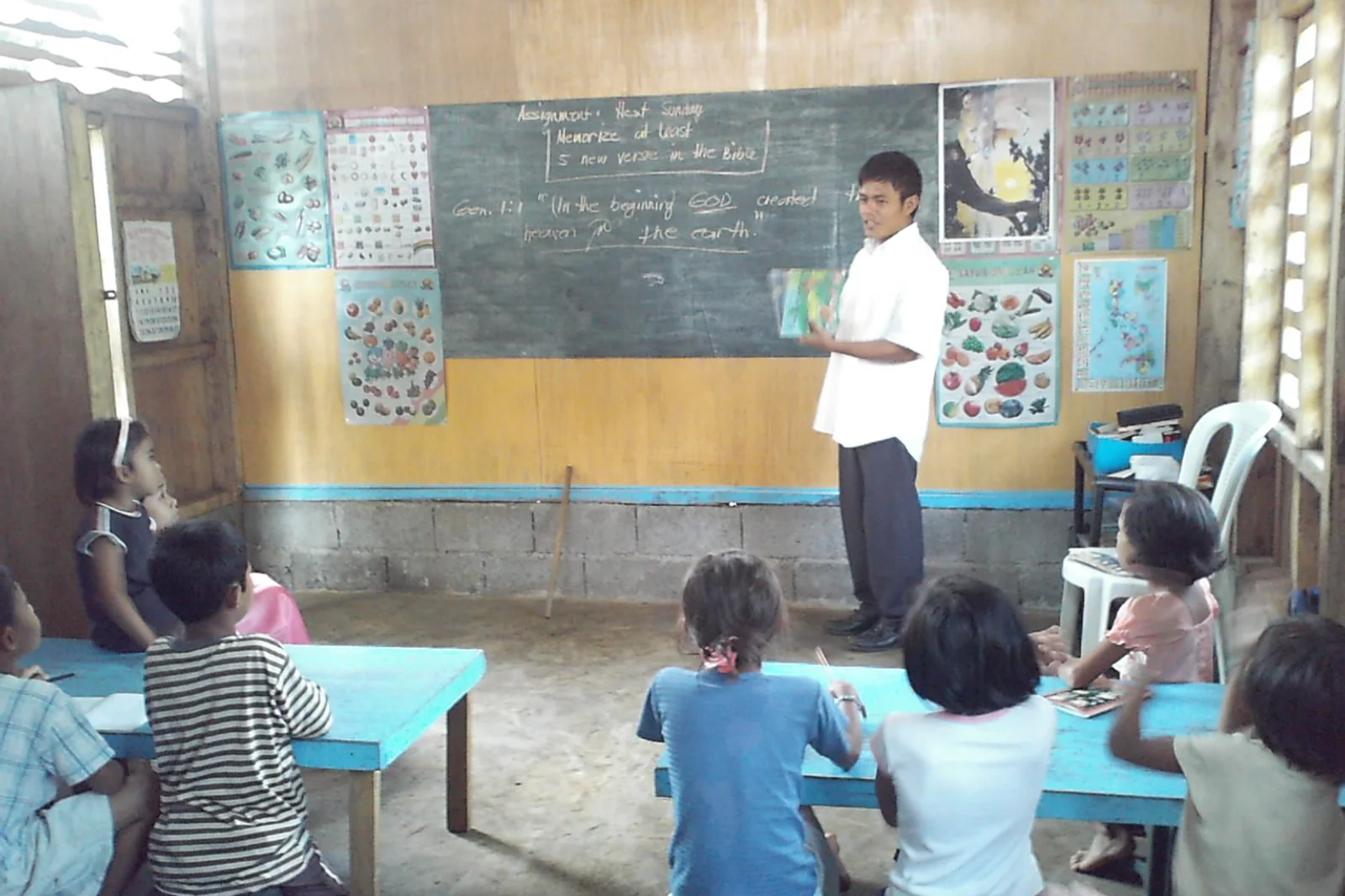 A classroom with a teacher standing at the front near a chalkboard, teaching young students who are seated at desks, with educational posters on the walls.