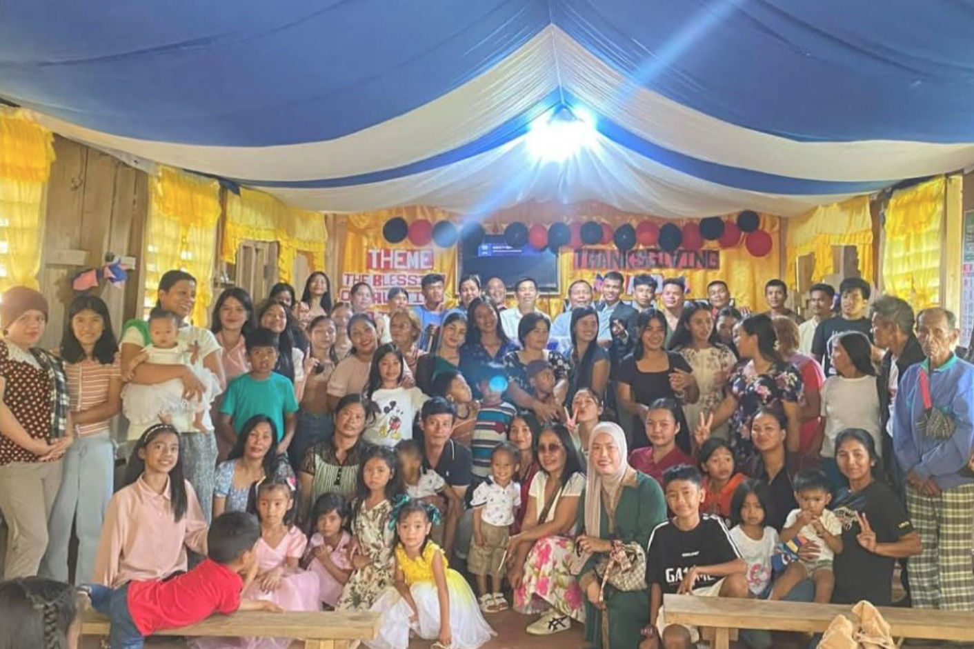 A large group of people, including children and adults, gathered inside a decorated community hall for a celebration. The hall has yellow curtains, blue and white drapes, and a banner in the background that reads 'Theme: The Blessing of...' with balloons and a TV screen overhead.
