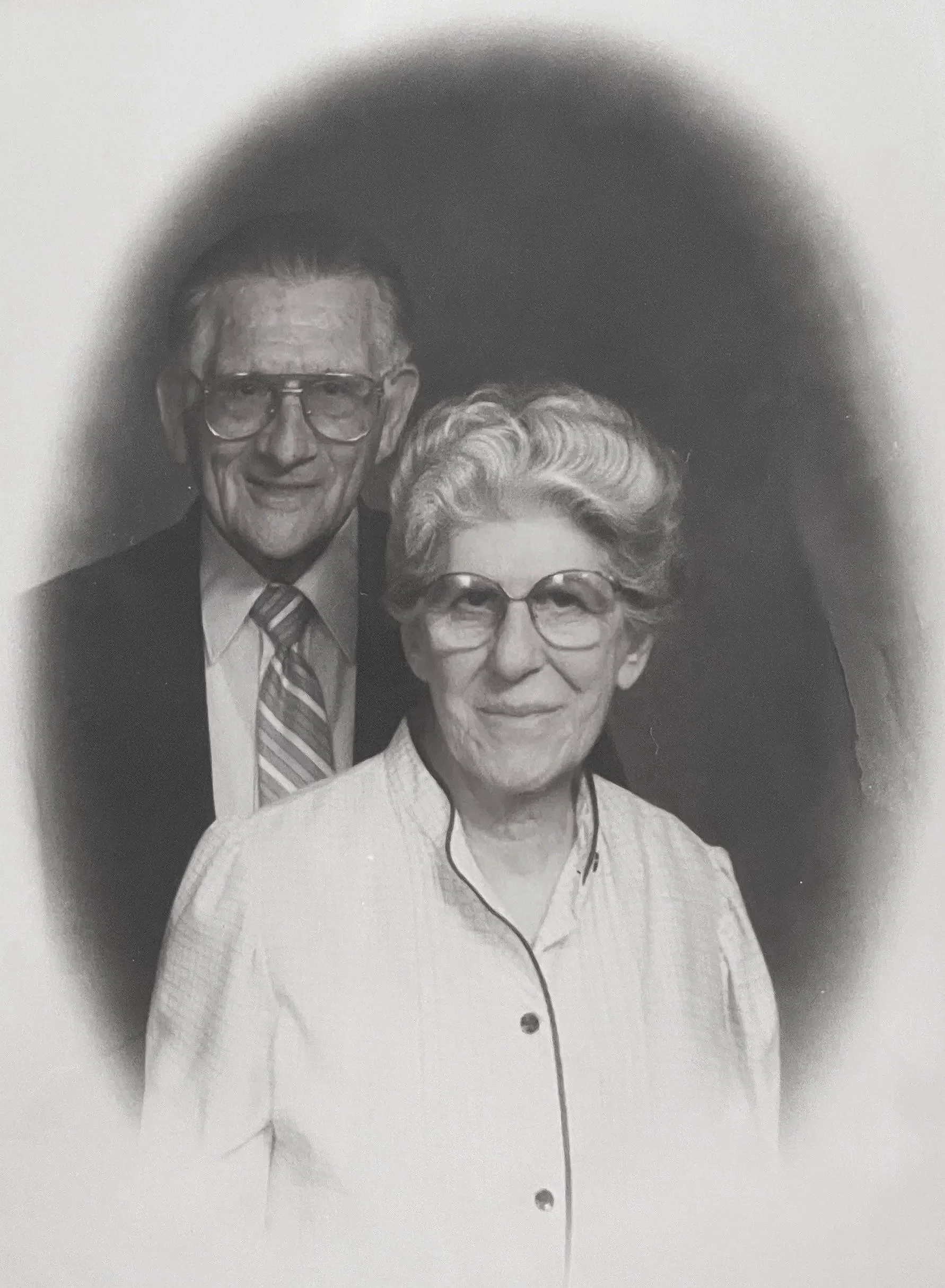 A black and white photograph of an elderly man and woman standing together, both wearing glasses. The man is dressed in a suit with a tie, and the woman is wearing a light-colored blouse or jacket.