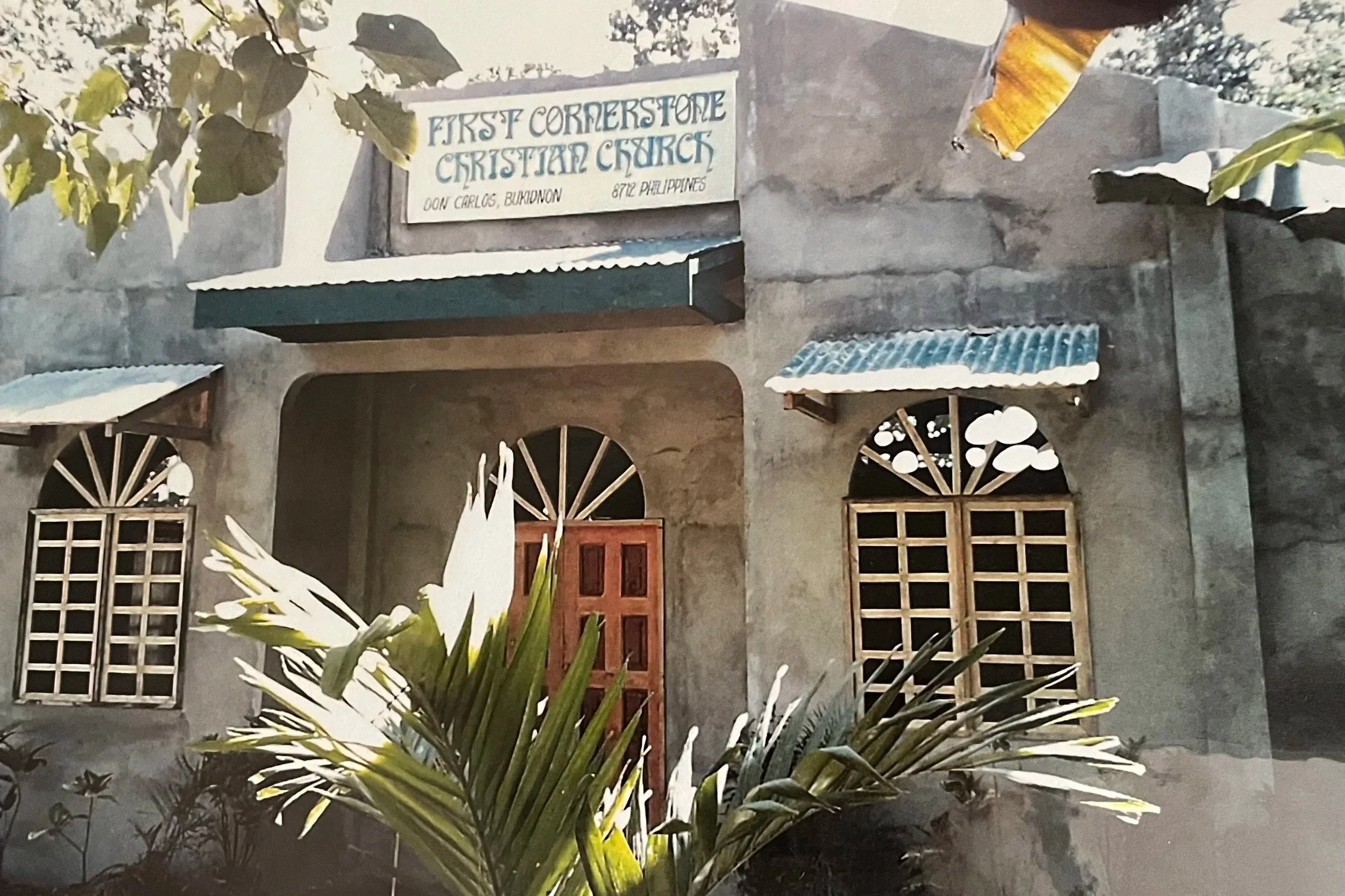 A building with a sign that reads "First Cornerstone Christian Church" located at Don Carlos, Bukidnon, Philippines. The building has two arched-framed windows with metal bars and a wooden door, with plants and trees surrounding it.