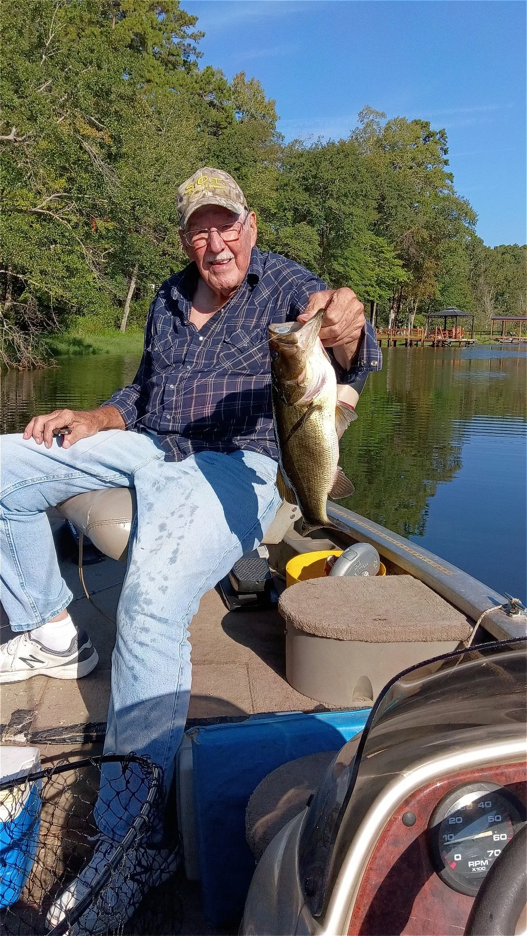 T-Bone's dad sits in a fishing boat holding up a freshly caught largemouth bass on a calm, tree-lined lake.