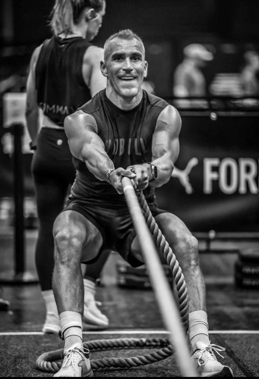 A shirtless man with a muscular build, holding a wooden stick and shouting aggressively in a gym setting, with gym equipment and bright lighting in the background.