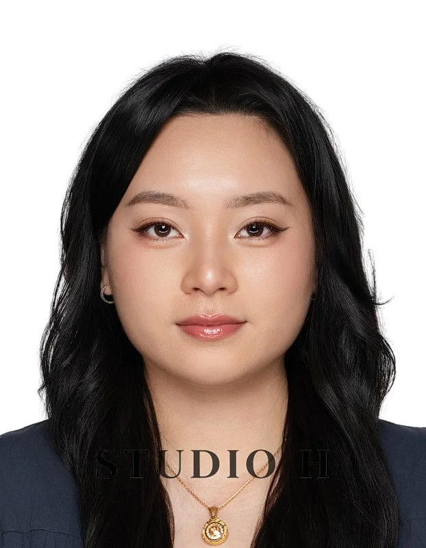 Headshot of a young Asian woman with long black hair, wearing a black top and a gold necklace with a pendant, against a white background.