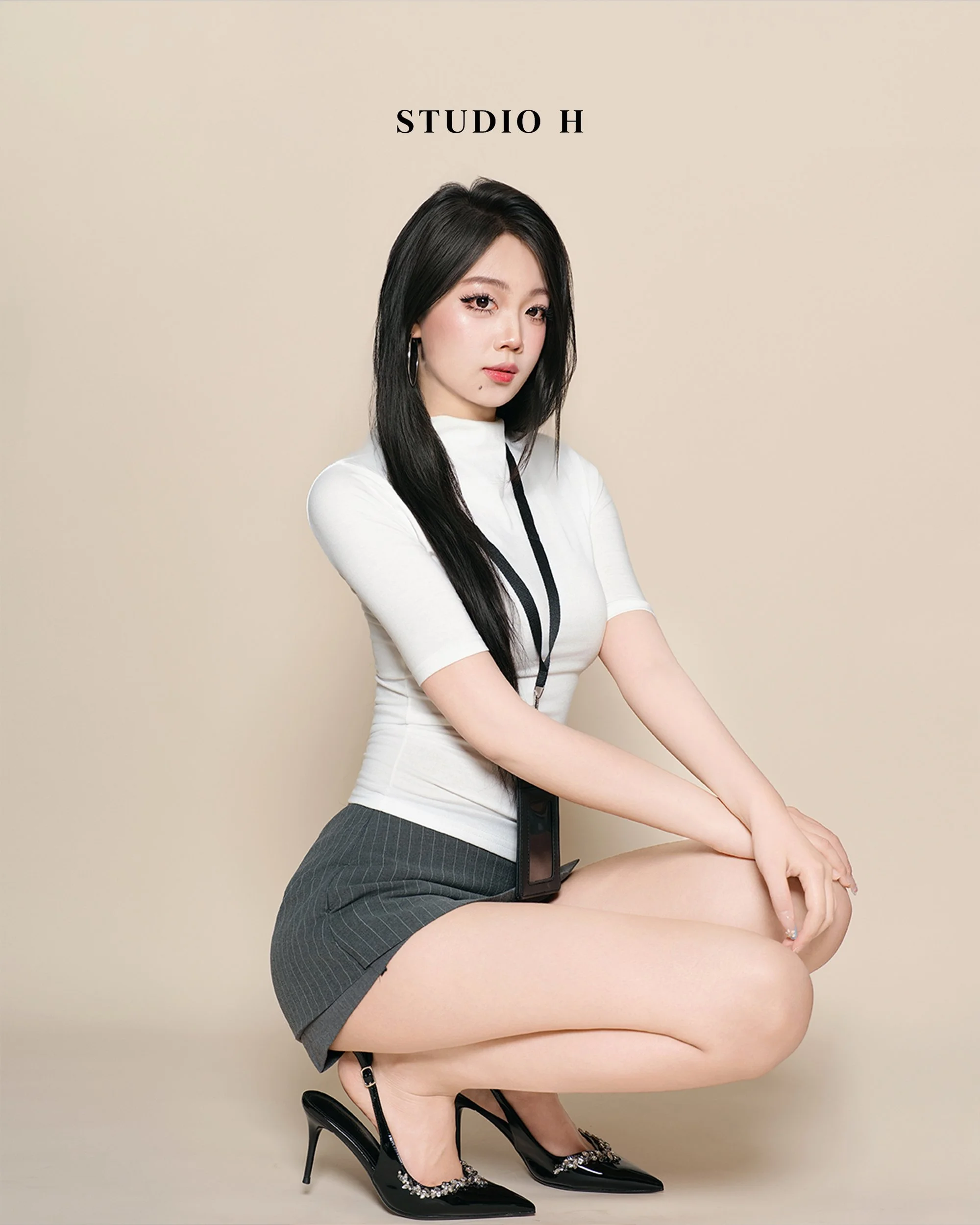 A young woman with long black hair, dressed in a white top and gray striped skirt, is crouching on the floor against a beige background with 'STUDIO H' written above her. She is wearing black high heels with decorative details and has a camera strap 
