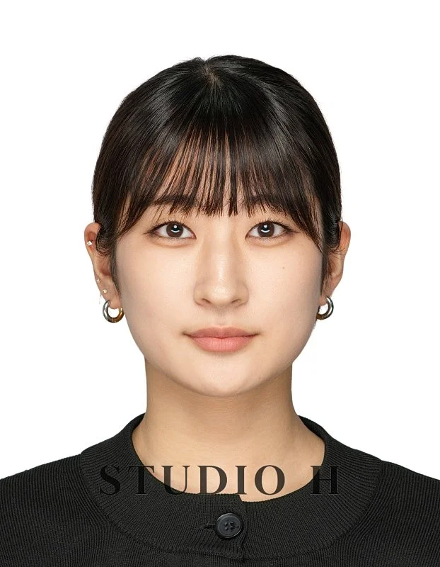 A young woman with short dark hair and bangs, wearing a black top and small earrings, looking directly at the camera with a neutral expression against a plain white background.