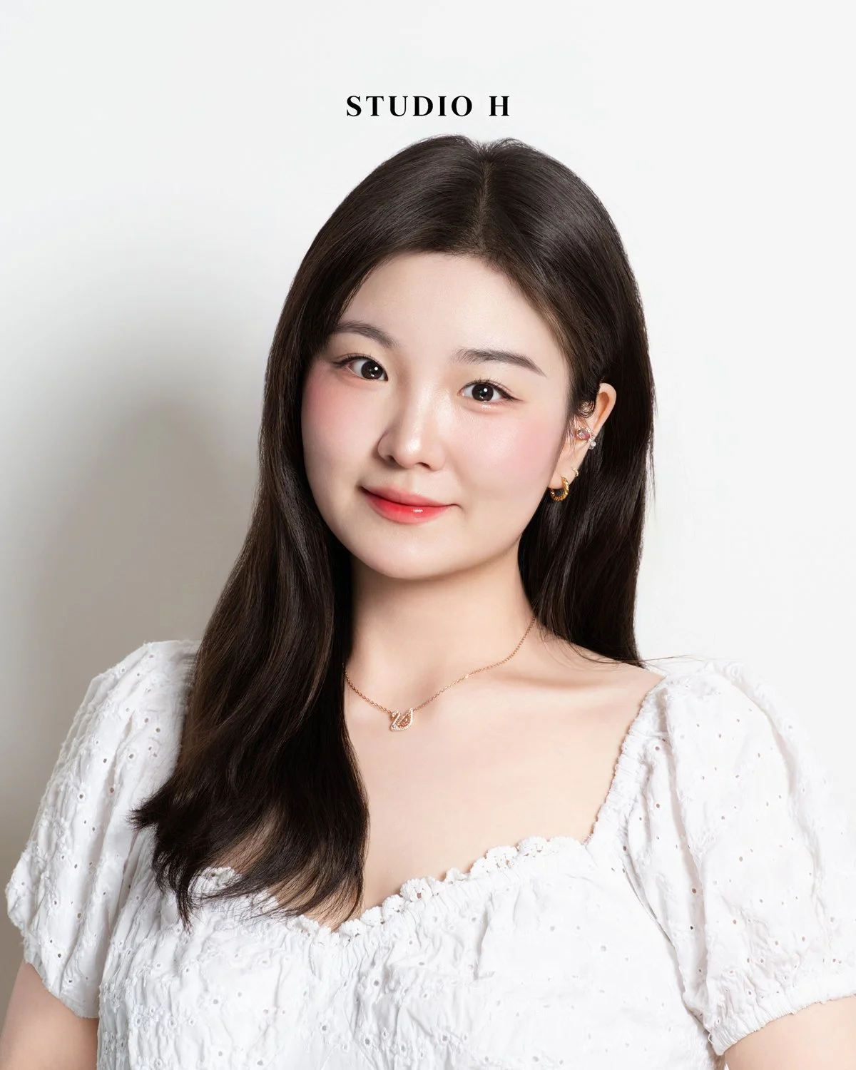 A young woman with long dark hair, wearing a white blouse with eyelet fabric, gold jewelry, and a necklace with a pendant standing against a light background with the text 'STUDIO H' above her head.