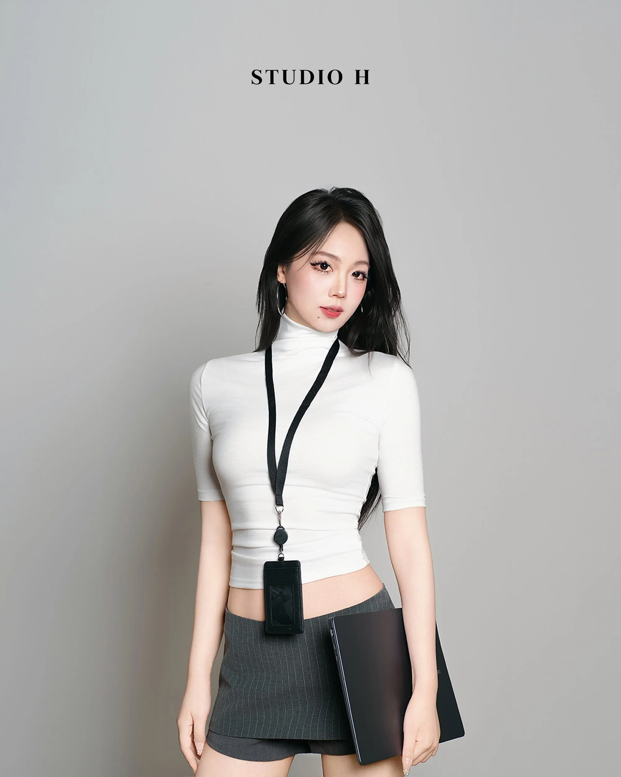 A young woman with long black hair, wearing a white turtleneck top and a dark gray pinstripe skirt, stands against a plain gray background. She has a black lanyard with a badge or ID around her neck and holds a black folder or tablet in her right hand. The text 'STUDIO H' appears above her.