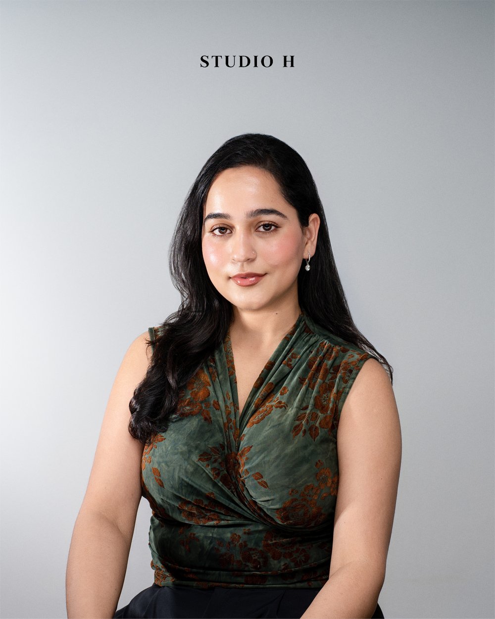 Portrait of a woman with long black hair, wearing a green and brown floral sleeveless top, sitting against a plain gray background with "STUDIO H" written above her.