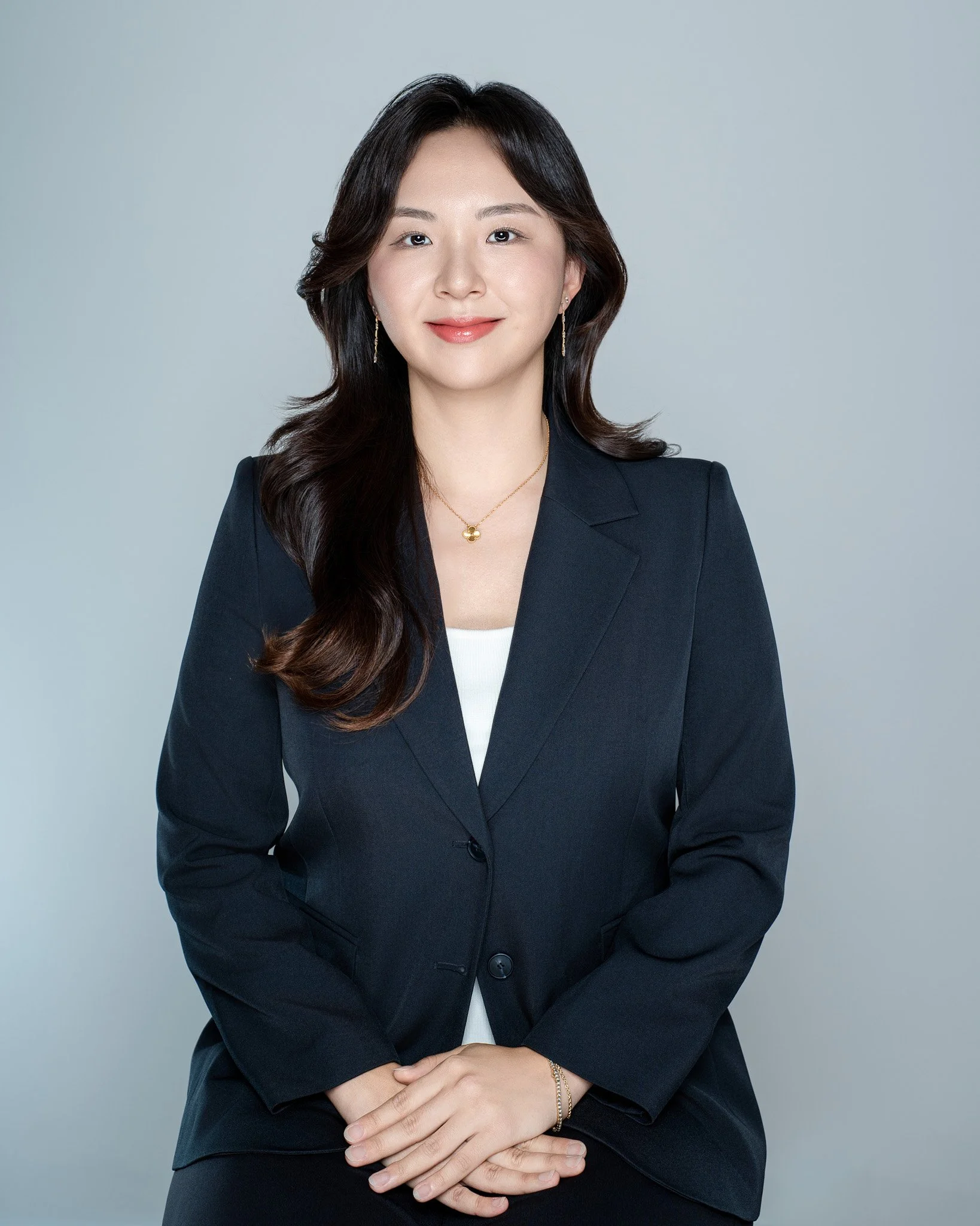 A woman with long wavy dark hair wearing a black blazer, white top, and gold jewelry, smiling in front of a gray background.