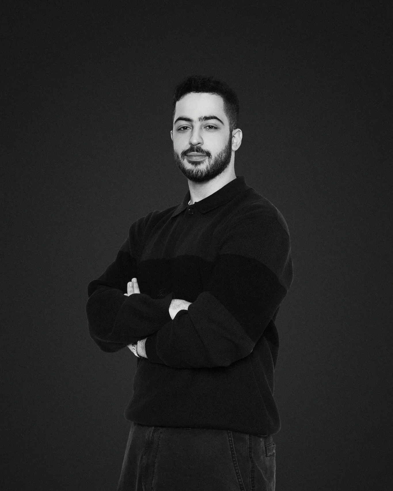Black and white portrait of a young man with a beard, wearing a dark long-sleeve shirt, standing with arms crossed against a plain dark background.