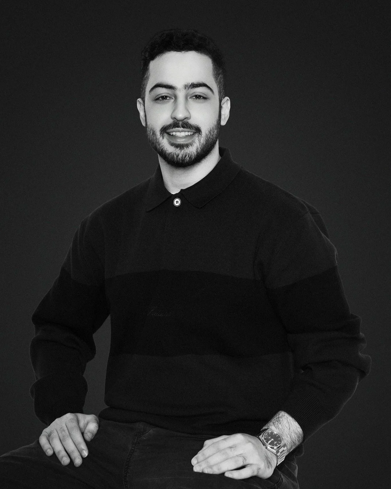 Black and white photo of a young man with dark hair, beard, and a watch, sitting against a dark background, smiling at the camera.