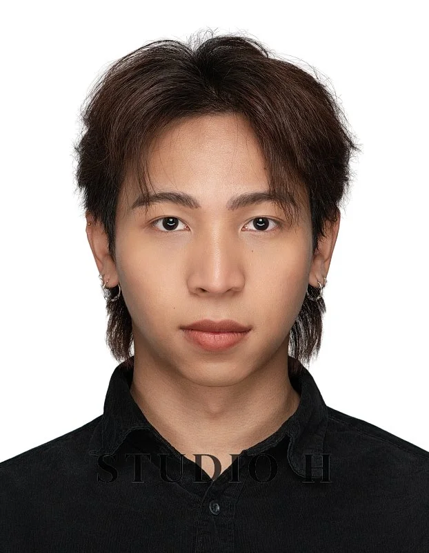 Portrait of a young man with medium-length brown hair, wearing earrings, and dressed in a black shirt, against a white background.