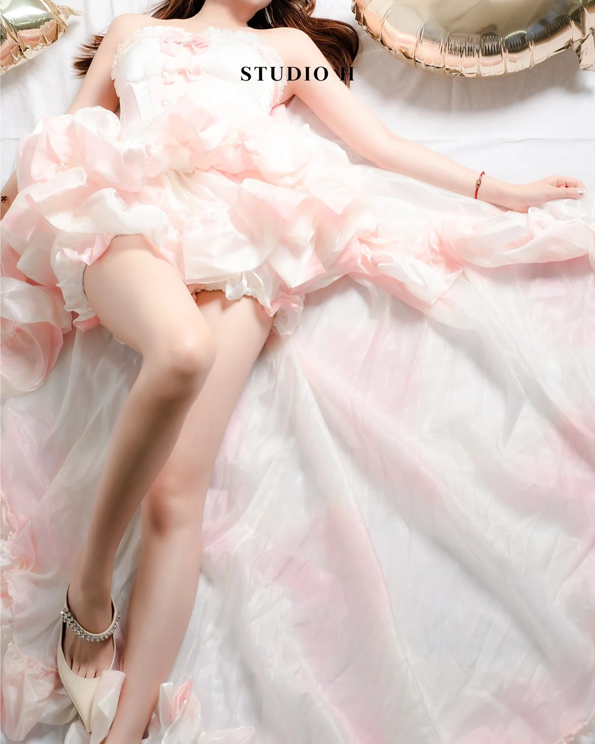A woman lying on a bed in a light pink, ruffled dress with gold high heels and a gold bracelet, with gold balloons in the background.