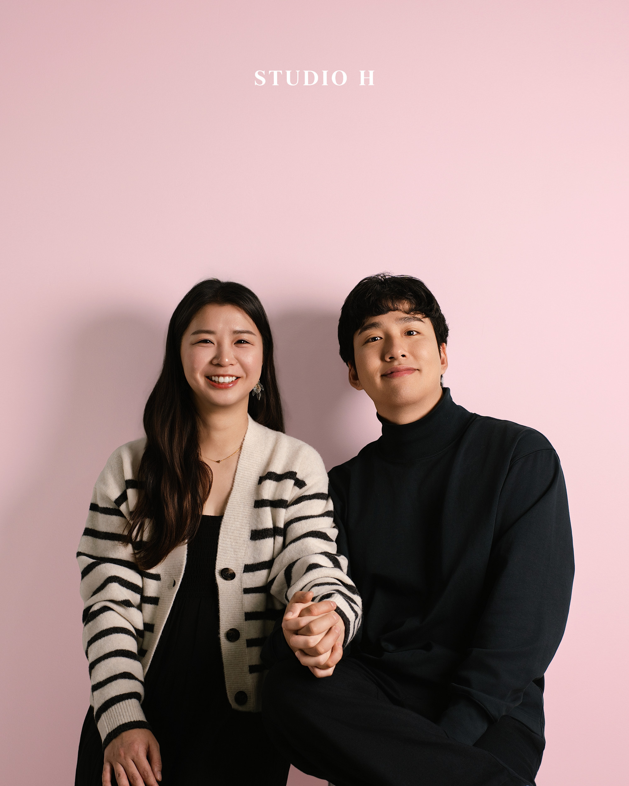 A woman and a man sitting together against a pink background, smiling and holding hands, with 'STUDIO H' written above them.