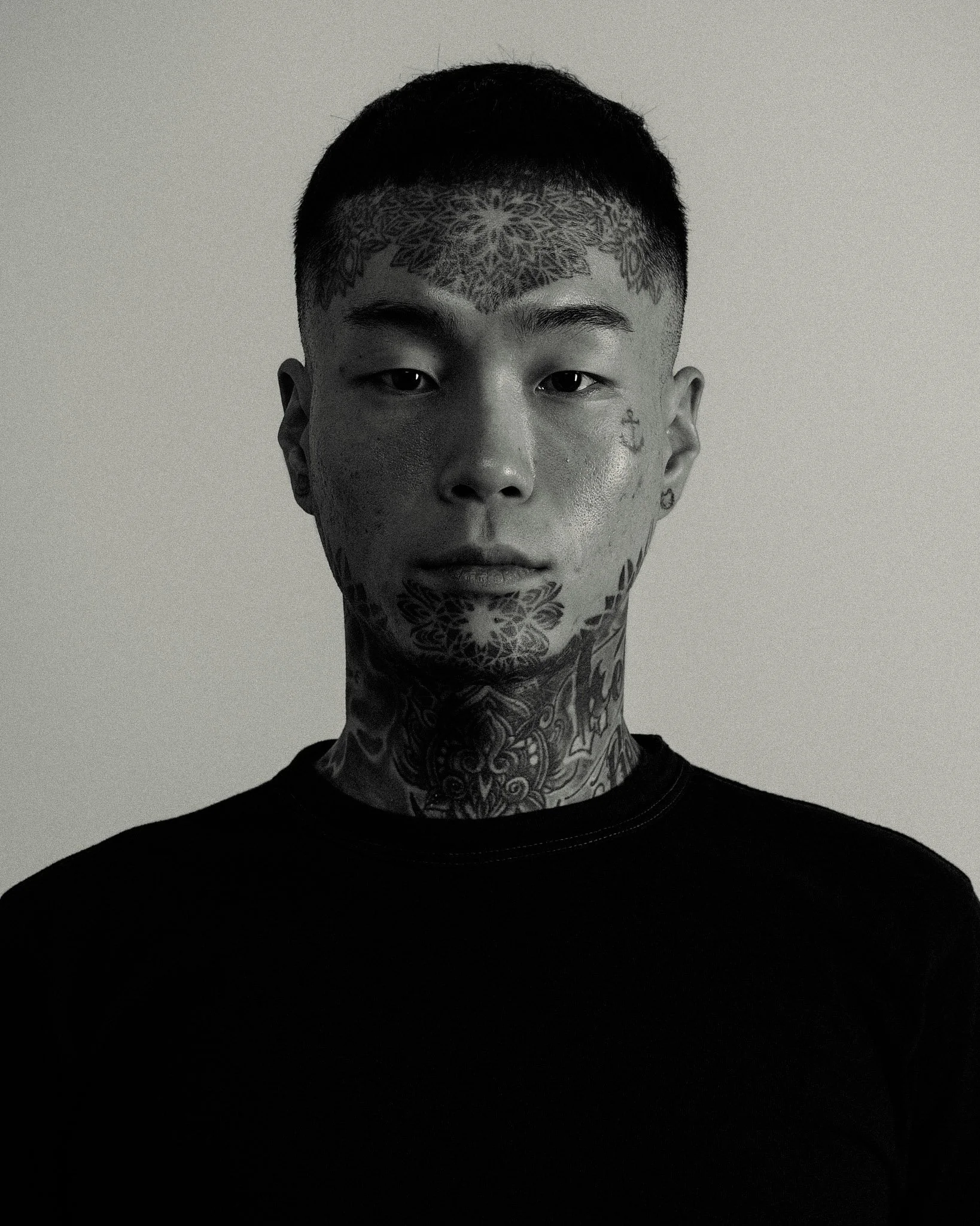 A black and white portrait of a young man with tattoos, including a large symmetrical design on his forehead and neck, and small tattoos on his face, wearing a black shirt and looking directly at the camera.
