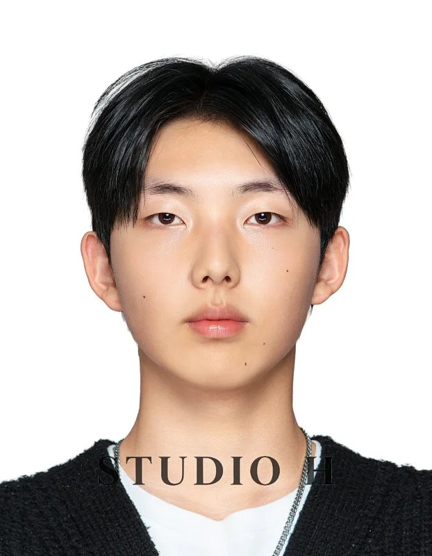 Portrait of a young man with black hair wearing a black sweater and a chain, against a white background, with the word "STUDIO" at the bottom.