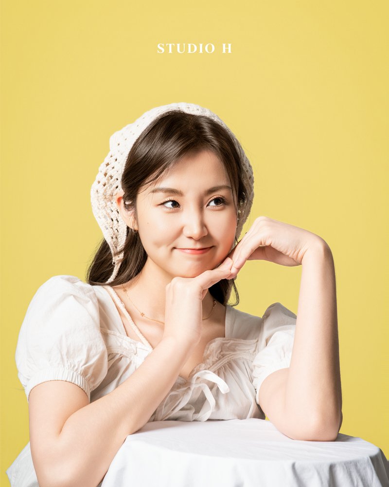 Young woman with dark brown hair, wearing a white knit hat and puff-sleeve blouse, resting her face on her hand with a playful expression, seated at a table against a yellow background with the text 'STUDIO H'.