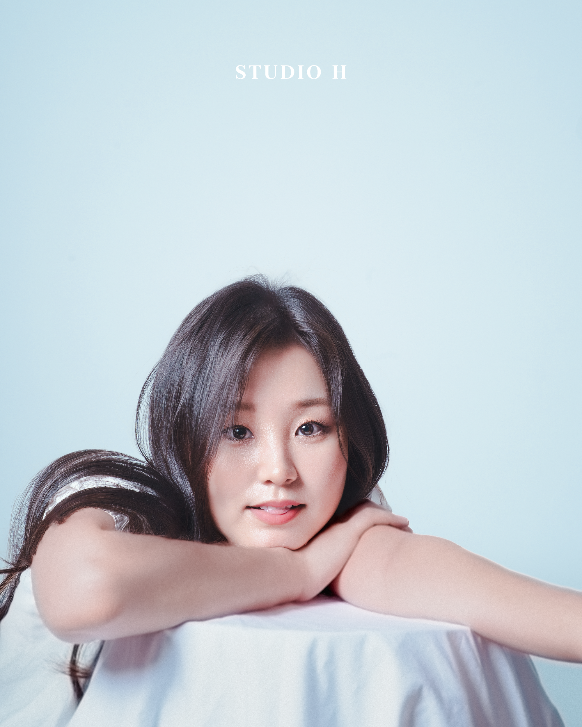 Portrait of a young woman with dark hair resting her head on her crossed arms, looking at the camera against a light blue background, with 'STUDIO H' text above.