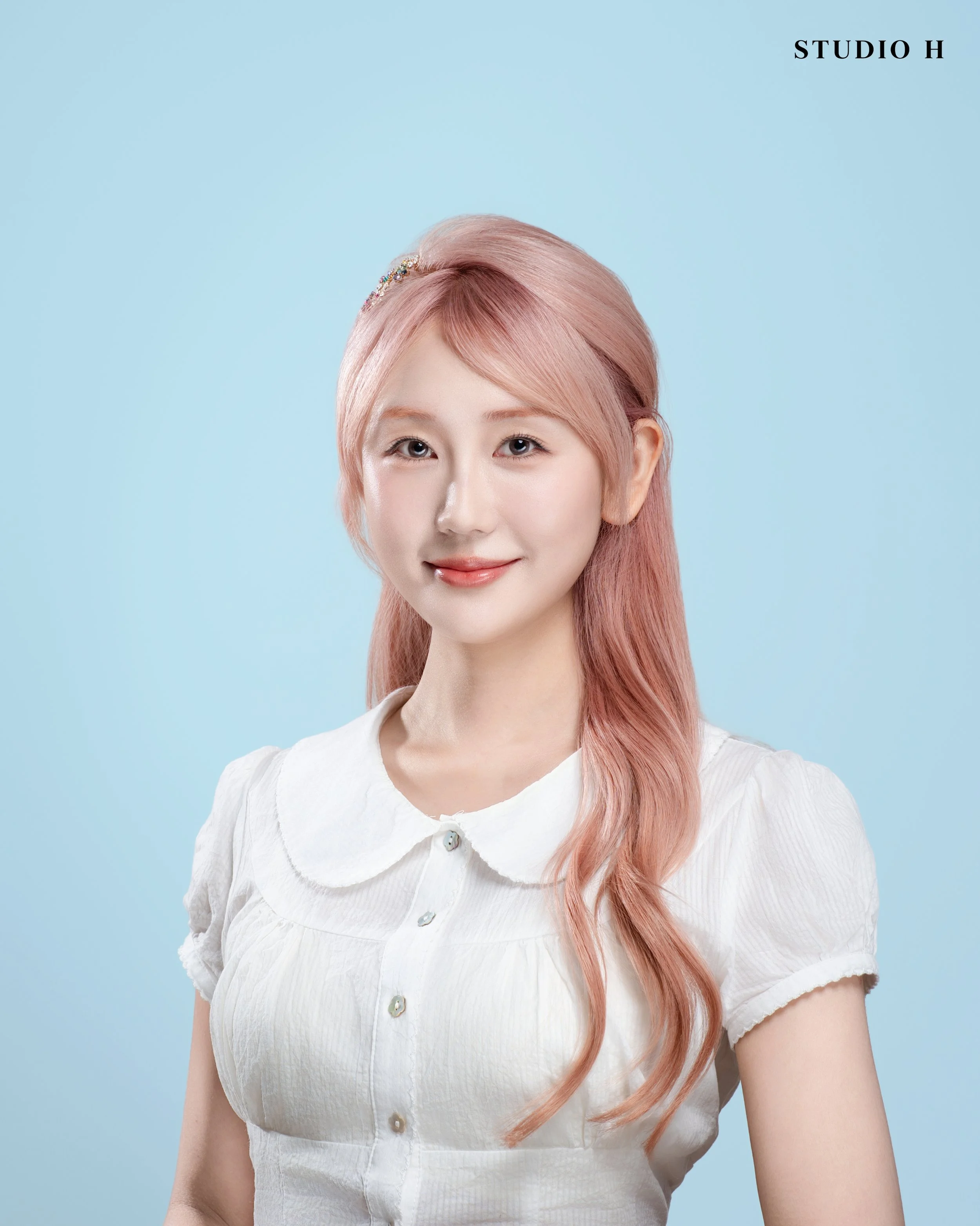A young woman with long, wavy pink hair, wearing a white blouse with a rounded collar, smiling softly against a light blue background. The text 'STUDIO H' is in the top right corner.