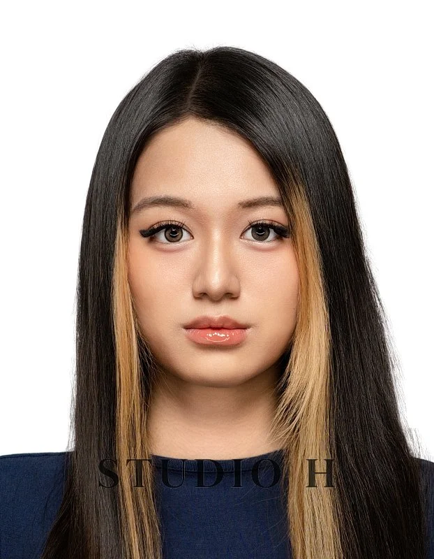 A close-up portrait of a young woman with long, straight dark hair, accented with blonde streaks, wearing a dark top, set against a plain white background.