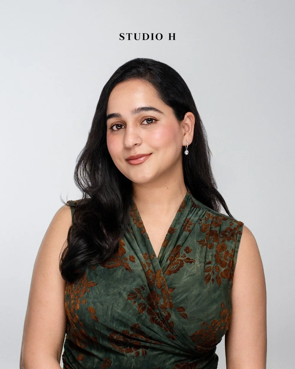 A woman with long dark hair, wearing a green floral sleeveless top, earrings, and a nose piercing, posing against a plain gray background with the text 'STUDIO H' above her.