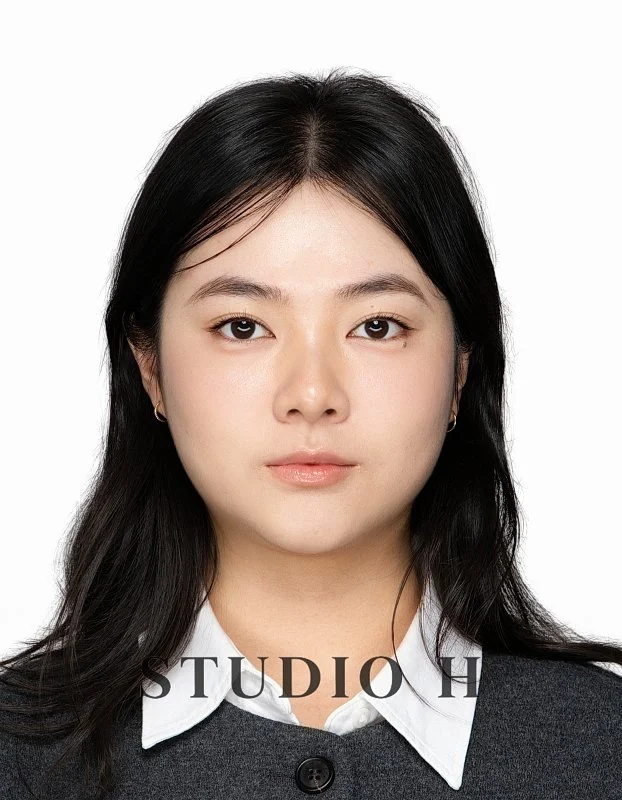 Young woman with dark hair wearing a white collared shirt and black sweater, looking directly at the camera against a plain white background.